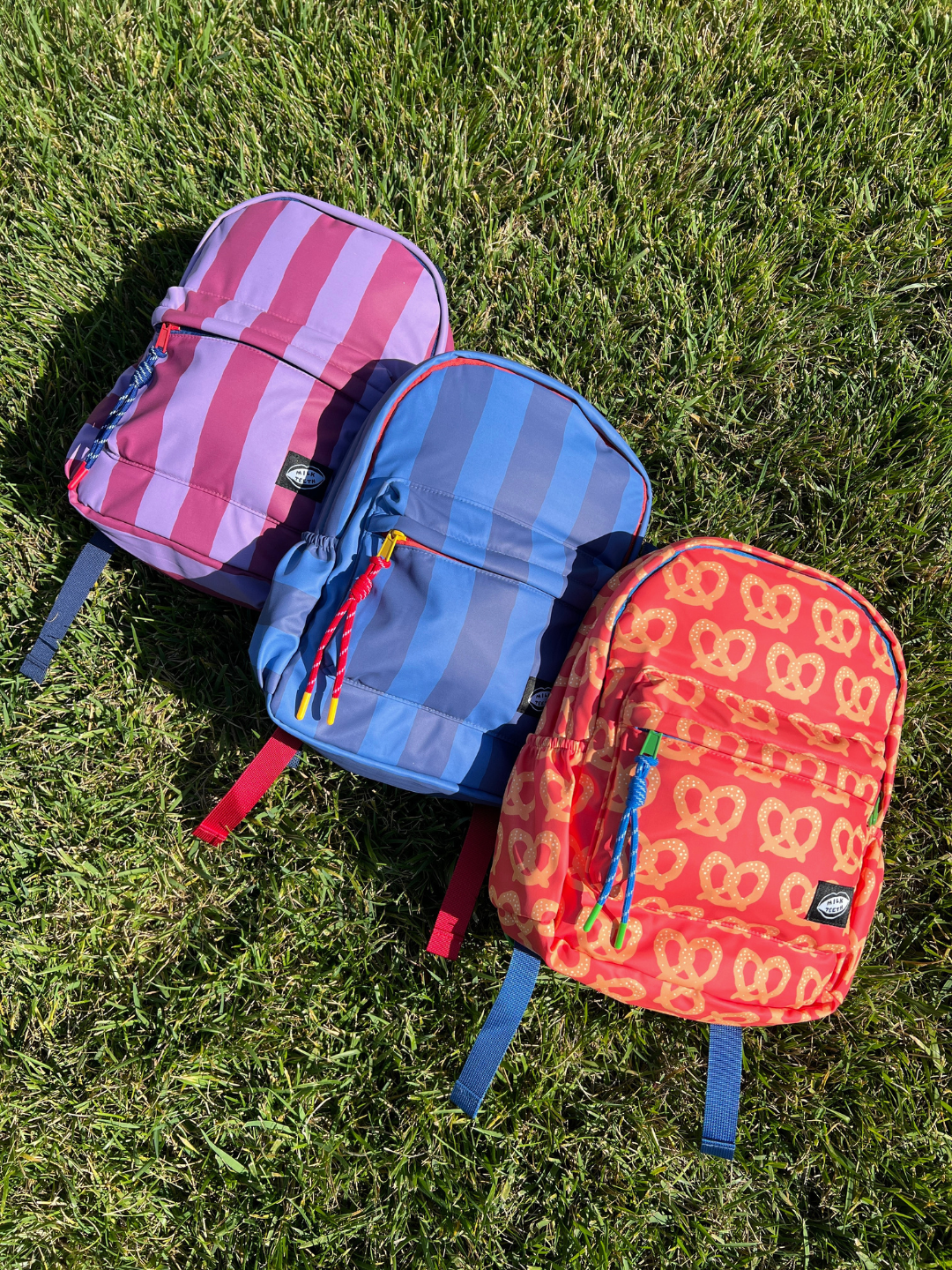 Three small colorful kids backpacks from Milk Teeth are arranged diagonally on a green grass lawn. The top backpack is maroon and purple with vertical stripes, the middle is blue with darker blue stripes, and the bottom is red-orange with a yellow pretzel pattern