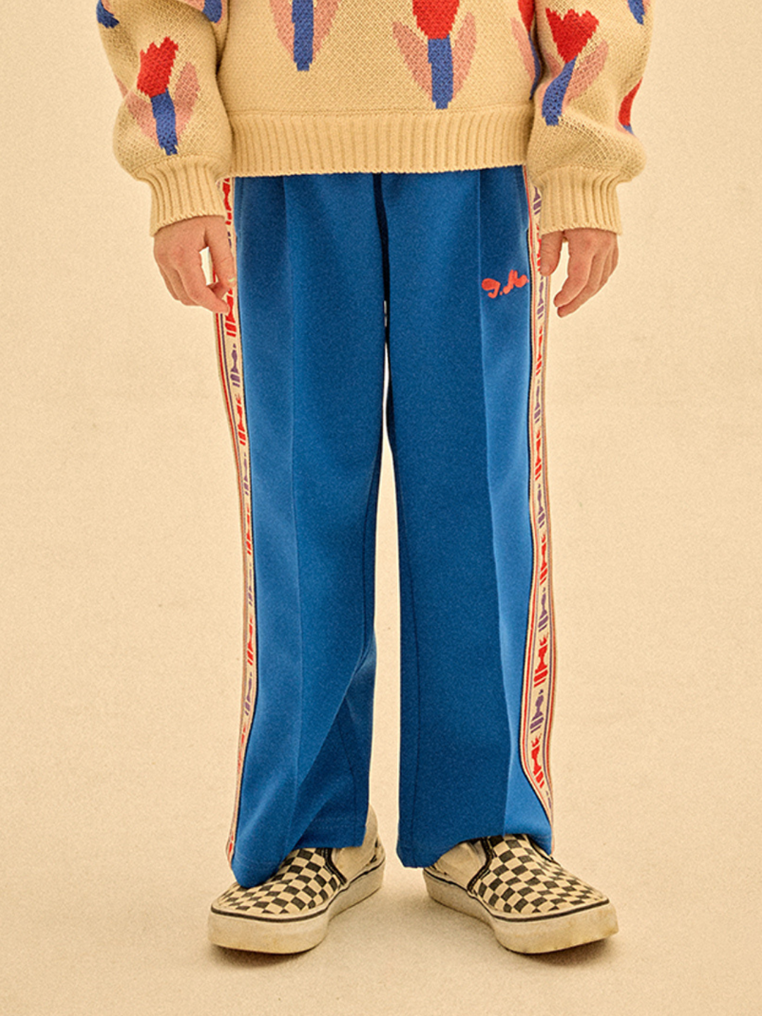 Blue | Wearing CHESS TRACK PANTS with side stripes, a patterned beige sweater, and checkered slip-ons, a person stands on a beige background, effortlessly showcasing style.