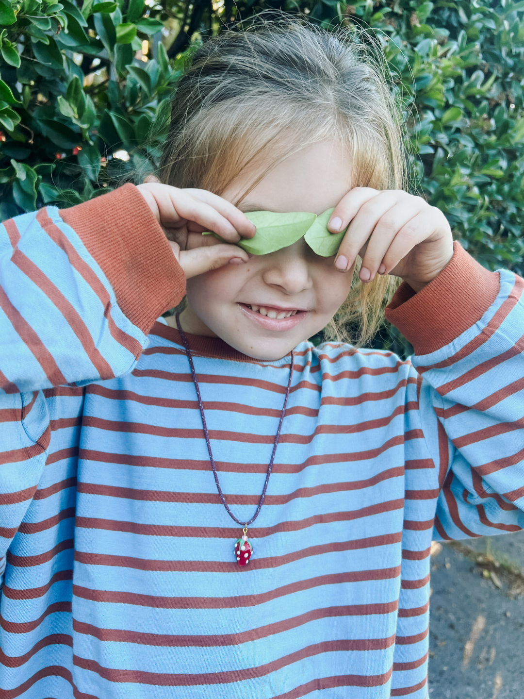 A child in a striped shirt holds green leaves over their eyes, standing by bushes, wearing an adjustable paracord bracelet with a handmade glass strawberry charm inspired by the Meyous jewelry FRAGOLA NECKLACE.