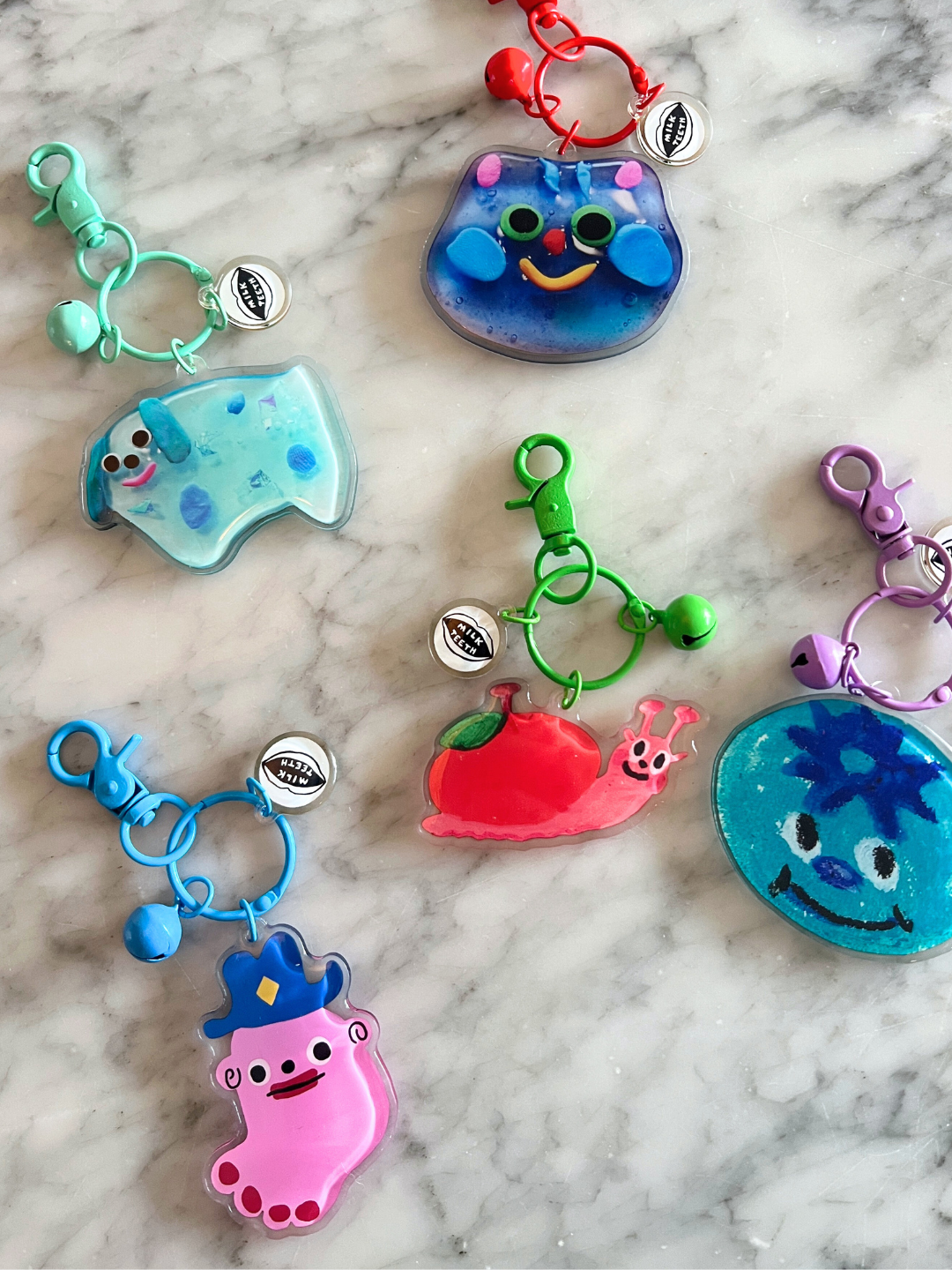 Snail | Five colorful SOUVENIR KEYCHAINs featuring Katie Kimmel's cartoon-style animal and character designs are displayed on a marble surface, each with a metal tag—perfect as playful charms or bag accessories.