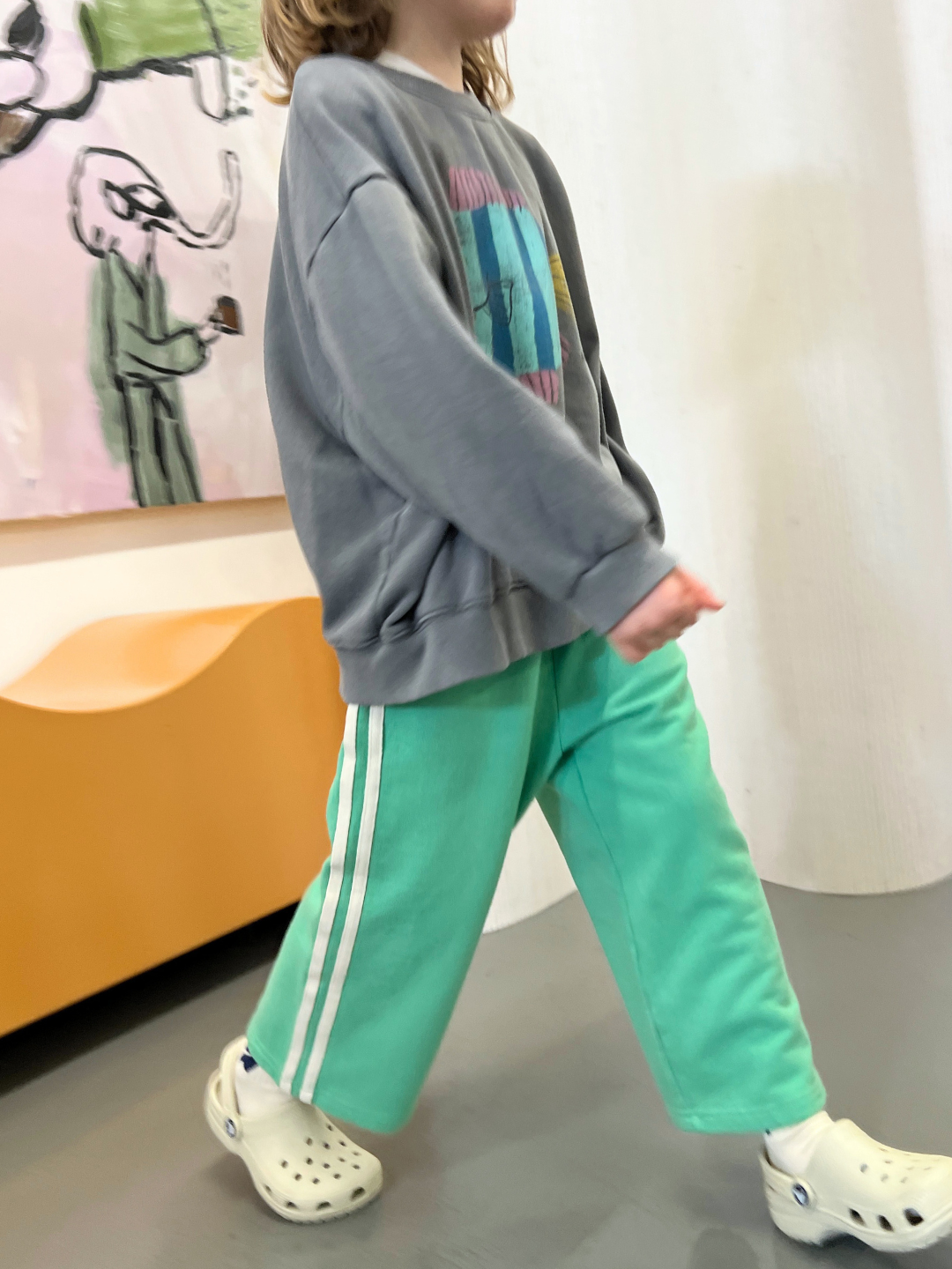 A child in a gray sweatshirt, Minty Stripe track pants, and white Crocs strolls indoors past wall art.