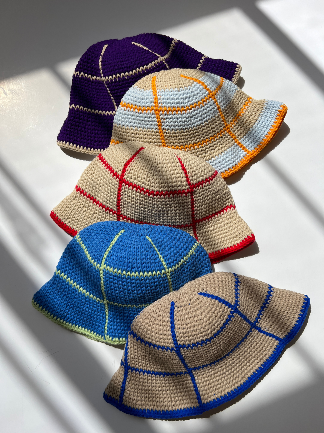 Five HAND-CROCHETED GRID HATS - 3-6Y are arranged in two rows under the sunlight, casting shadows. Made from cotton yarn, the hats come in various colors and patterns, including purple with white, yellow with blue and orange, tan with red, blue with green, and tan with blue.