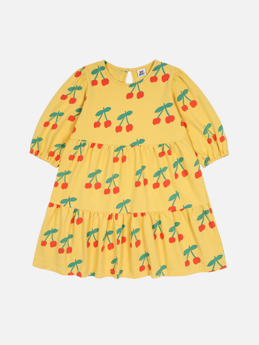 Image of The CHERRY RUFFLE DRESS is a yellow children's dress with three-quarter sleeves, a tiered skirt, and a cherry pattern featuring red cherries and green stems on a white background.