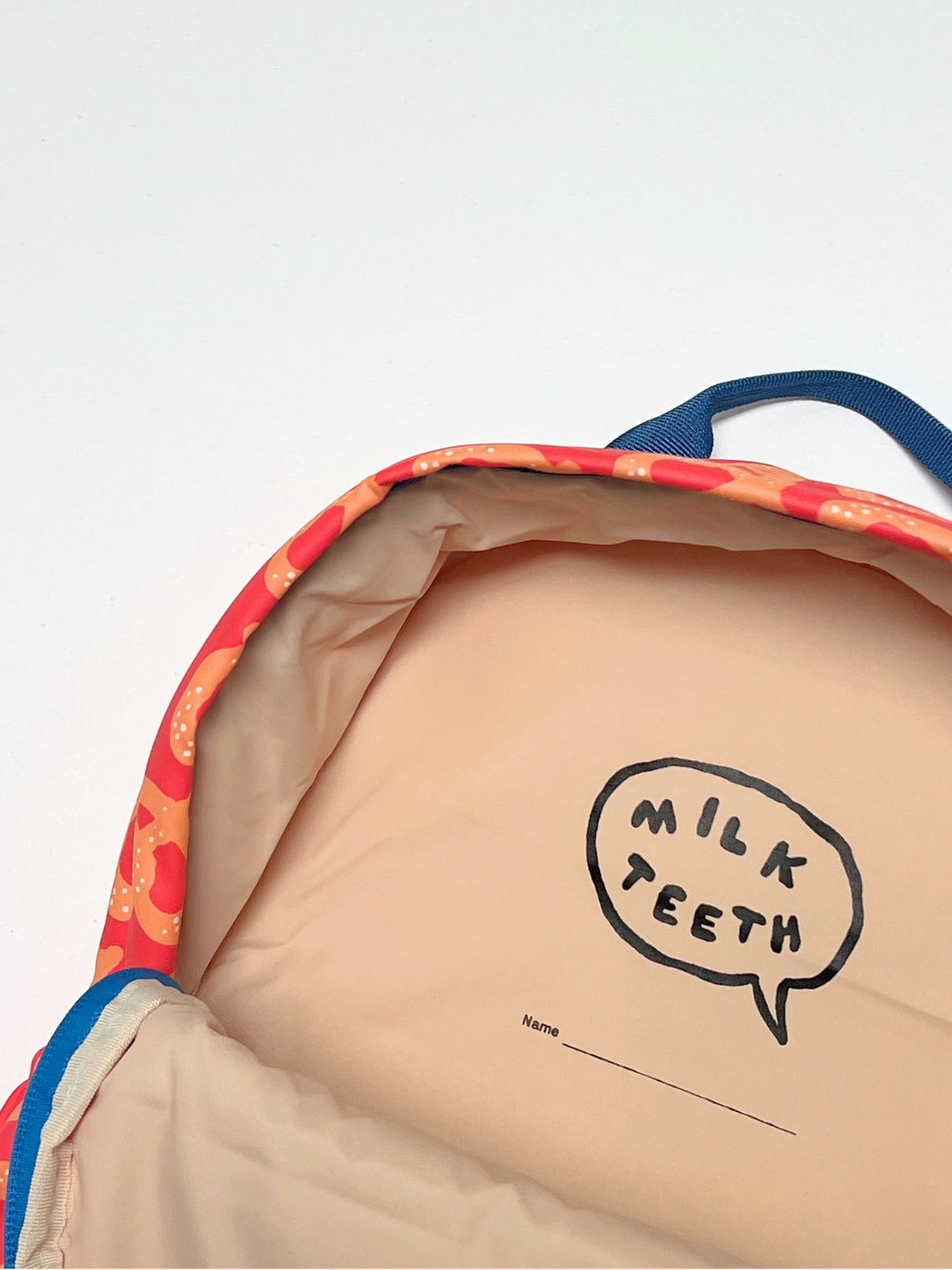 Red Pretzel | Close-up of the inside of a puffy quilted backpack with a beige interior, red outer, blue handle, and a printed speech-bubble logo reading 'MILK TEETH' on the lining with a name label space.