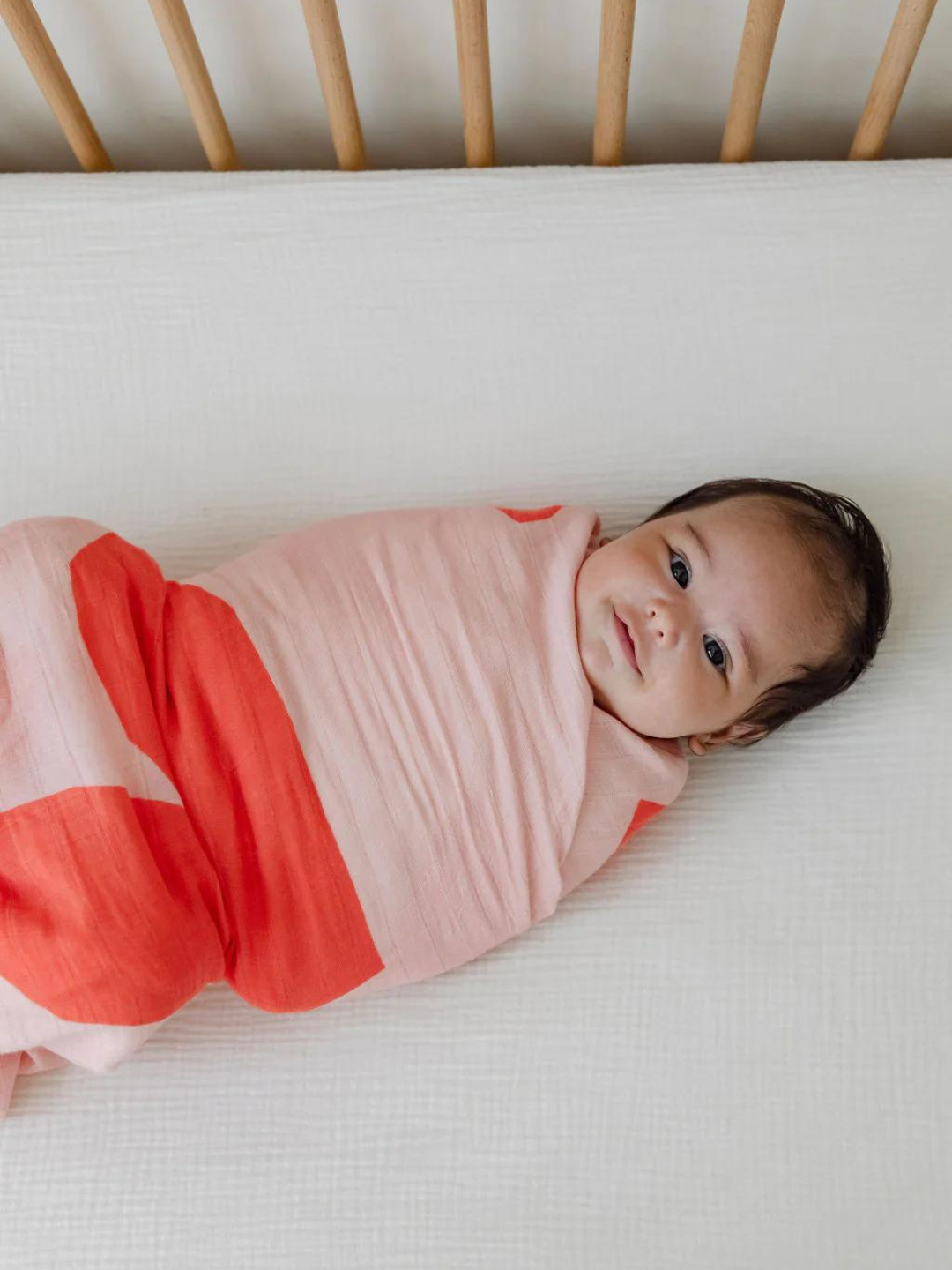 A baby wrapped in a pink and red ORGANIC BAMBOO SWADDLE WRAP lies on a white surface, looking up.