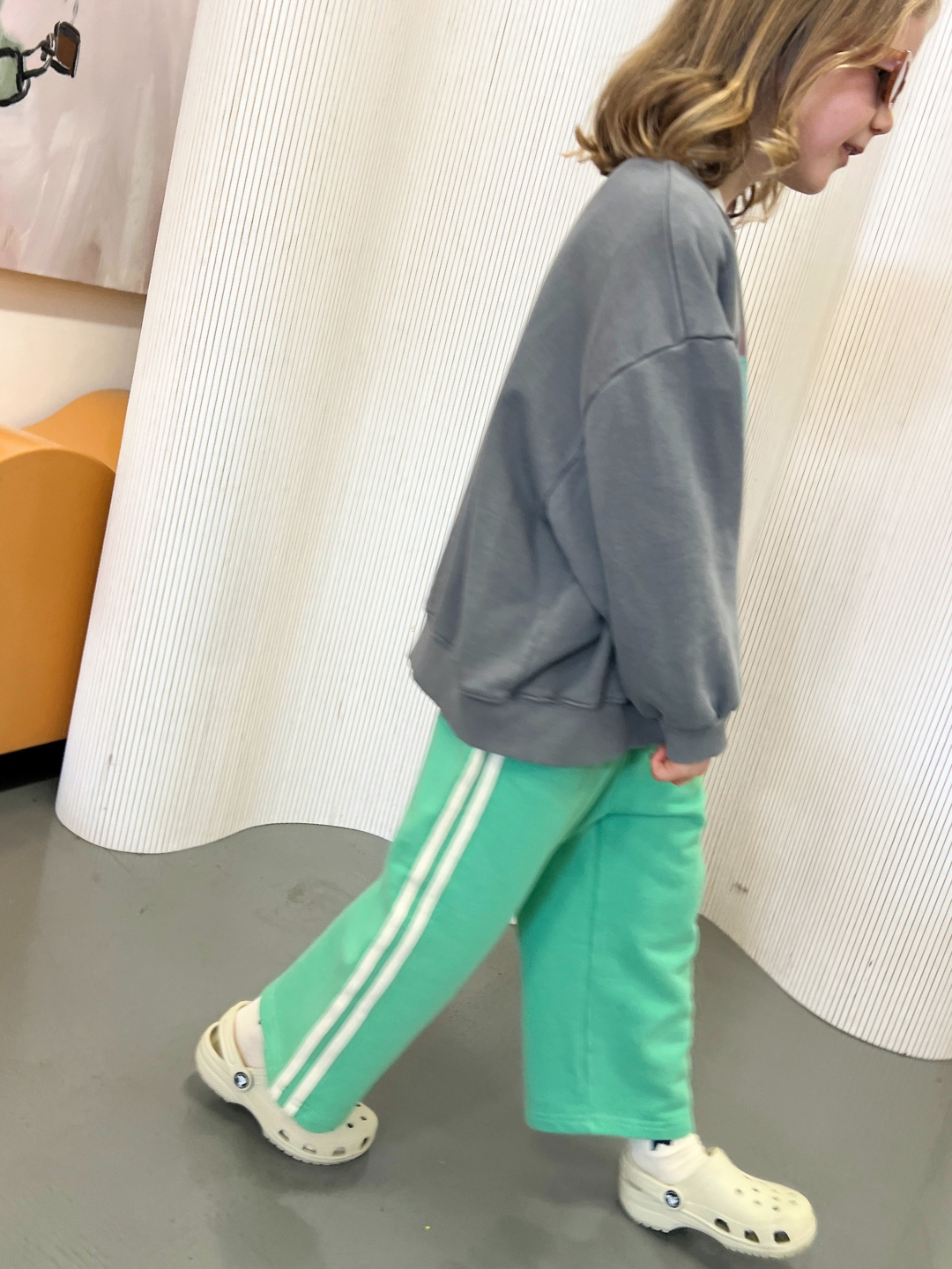 A child in a gray sweater and MINTY STRIPE TRACK PANTS from Bon Bon Butik, wearing light-colored Crocs, strolls indoors beside a white textured wall.