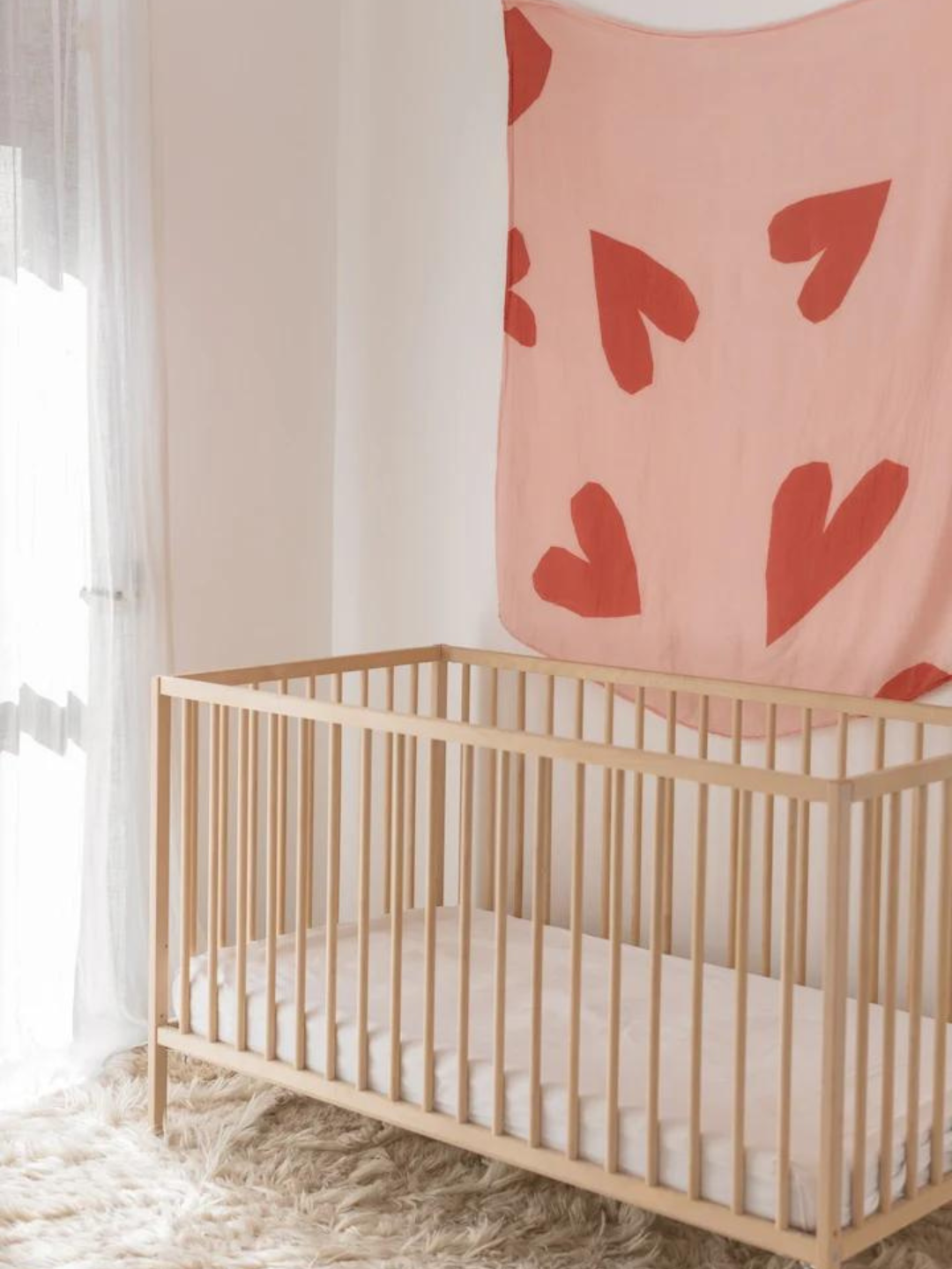 A wooden crib with a white mattress is placed in a room with a fluffy carpet; a pink blanket with red heart designs hangs on the wall behind the crib, alongside some delicate ORGANIC BAMBOO SWADDLE WRAPS.