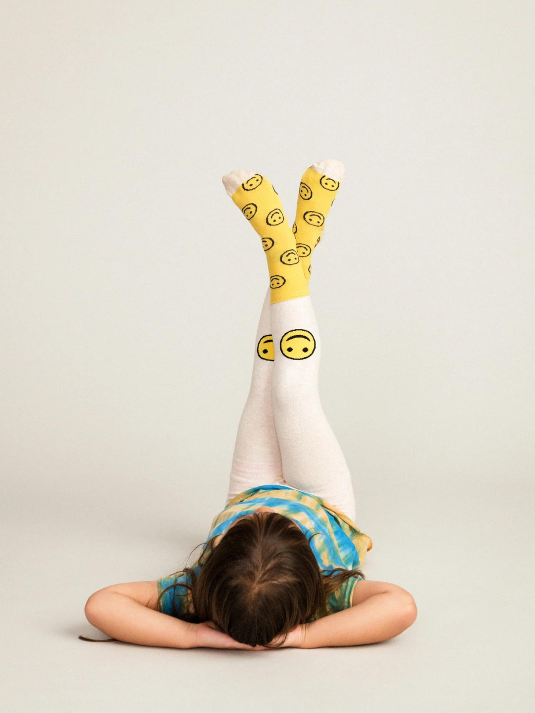 A child with her legs crossed in the air, wearing a pair of kids' leggings in cream with yellow smileys at the knee, together with matching socks