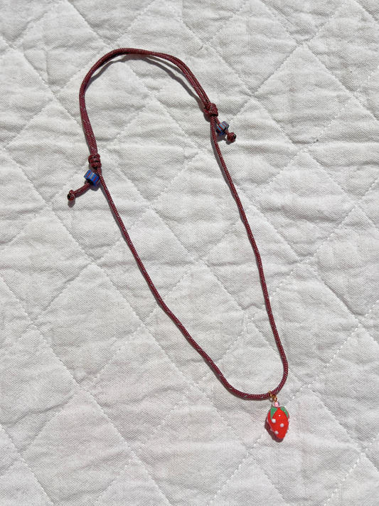 Image of The FRAGOLA NECKLACE by Meyous features a red cord with knotted ends and a handmade glass strawberry charm, shown on quilted white fabric.