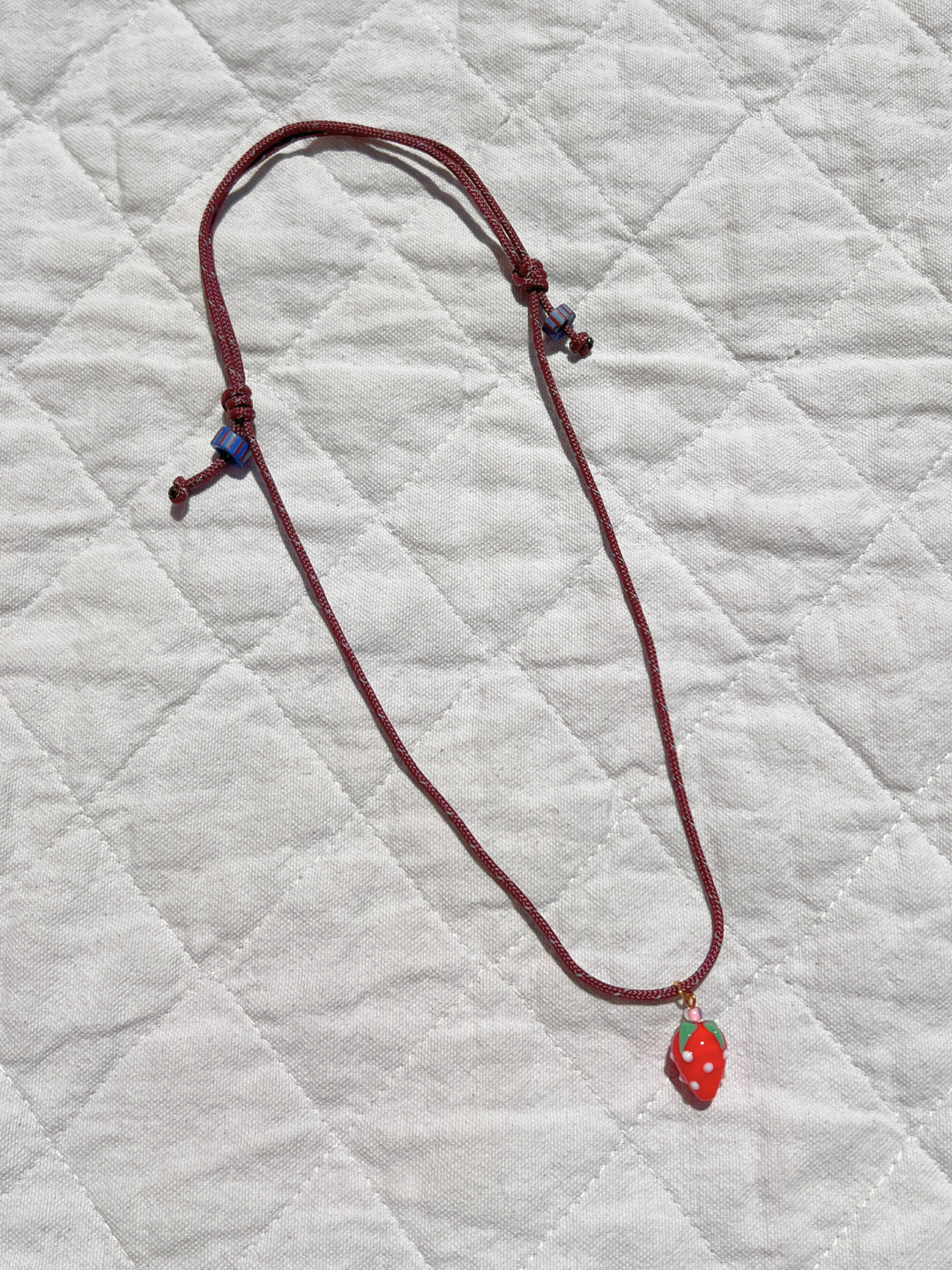 The FRAGOLA NECKLACE by Meyous features a red cord with knotted ends and a handmade glass strawberry charm, shown on quilted white fabric.