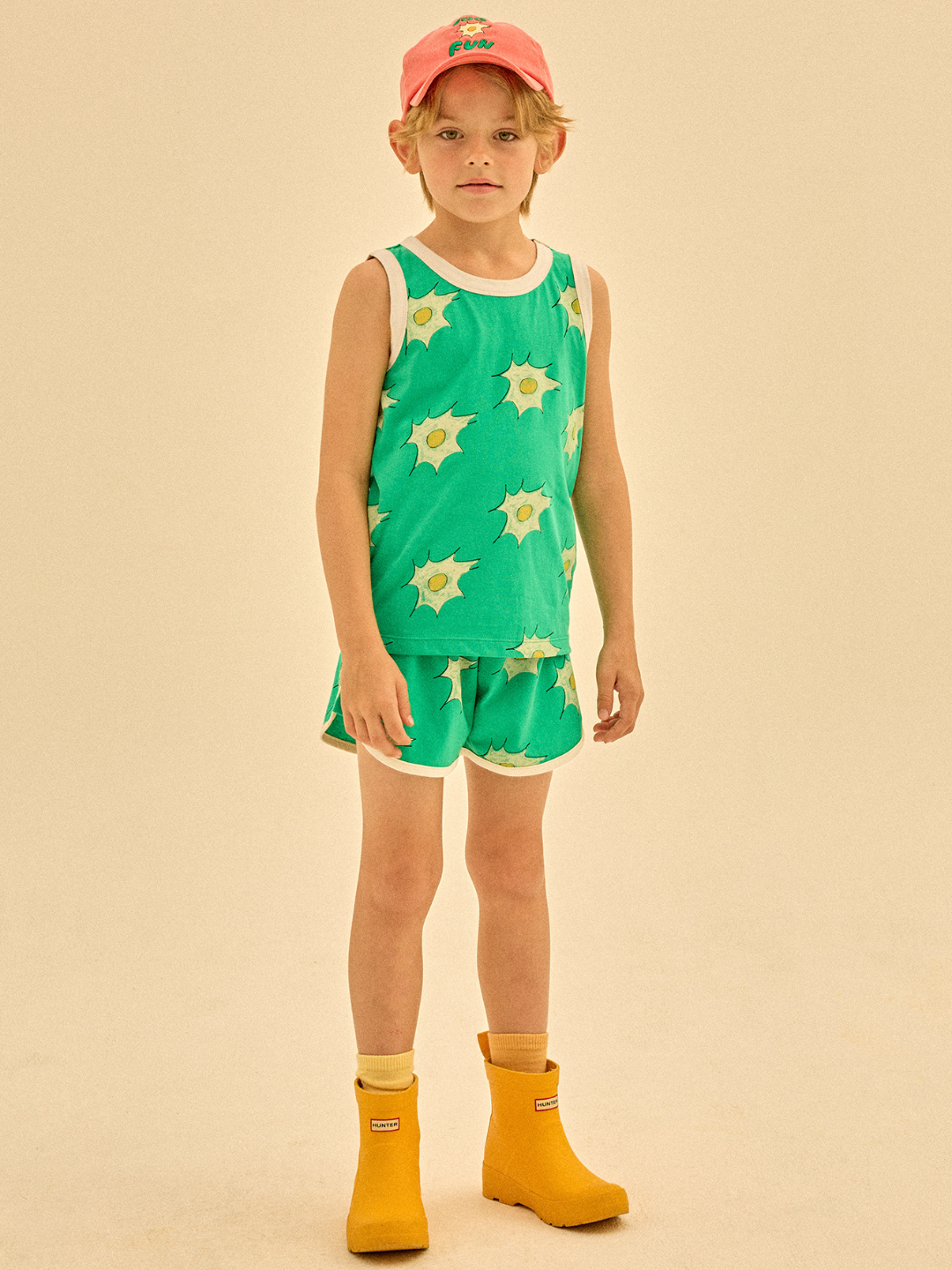 A child wearing the EGG SUMMER SET, consisting of a matching green tank top and shorts with star patterns, is accessorized with a pink cap and yellow boots, standing against a plain background.