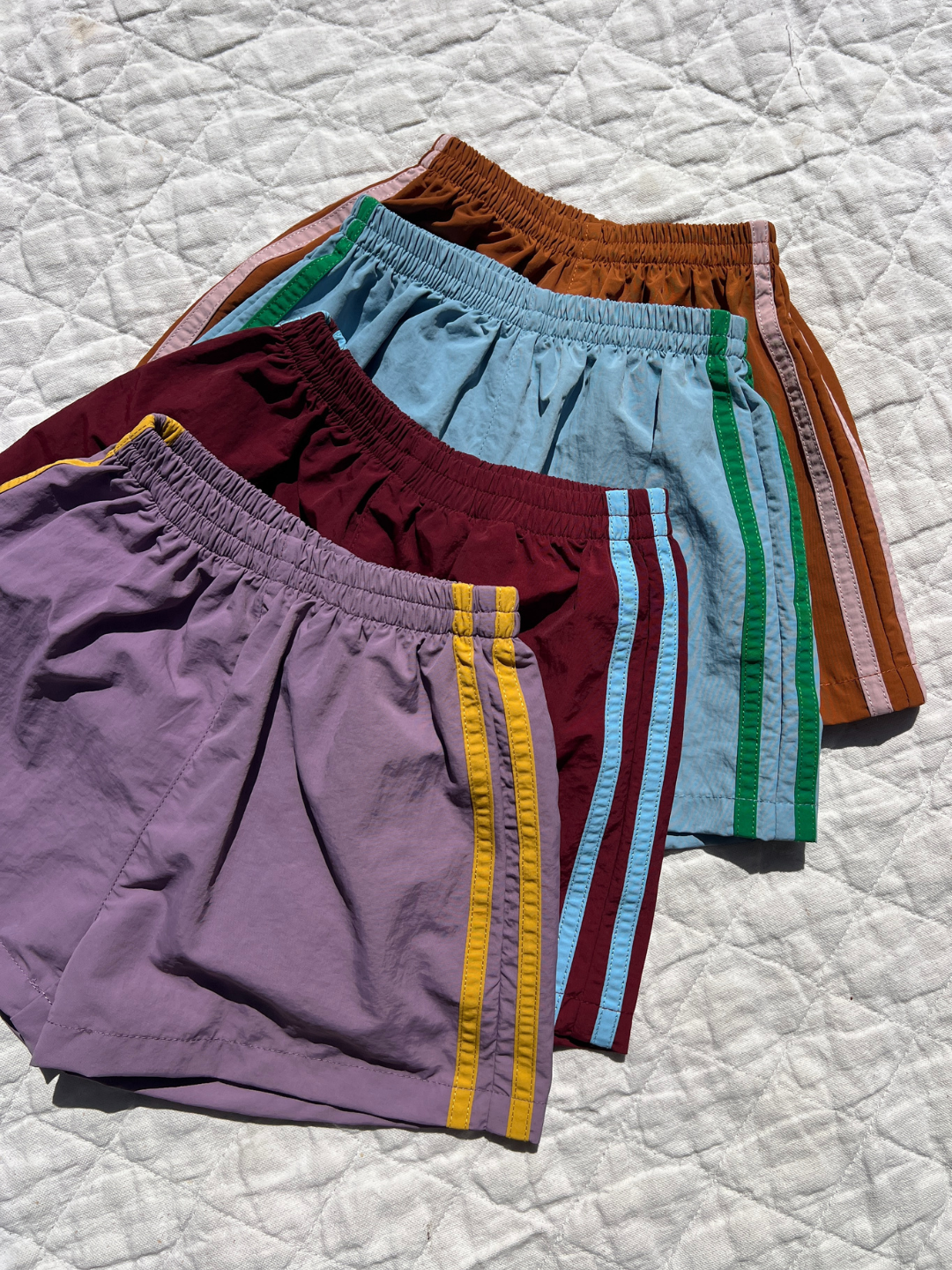 Four pairs of SPRINTER SHORTS with colorful stripes are arranged overlapping on a quilted white surface.