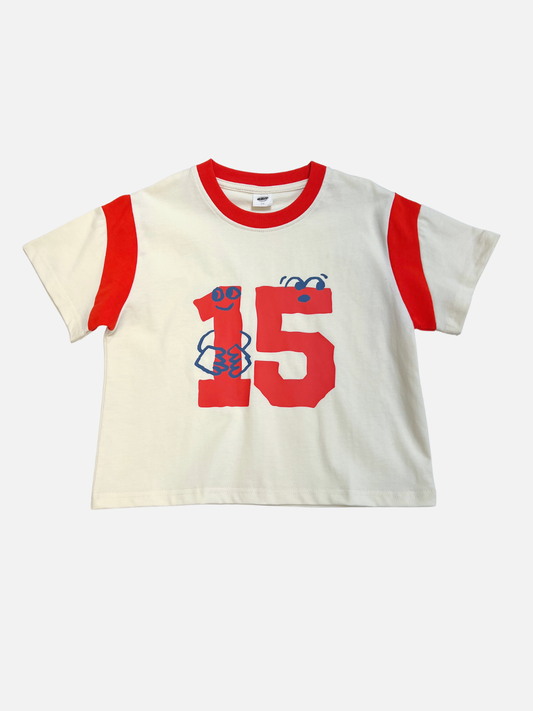 Image of The VARSITY TEE is a white cotton t-shirt with red sleeve stripes and collar, a bold red number 15, and a small blue graphic on the front. It features an oversized boxy fit for a classic sports jersey look.