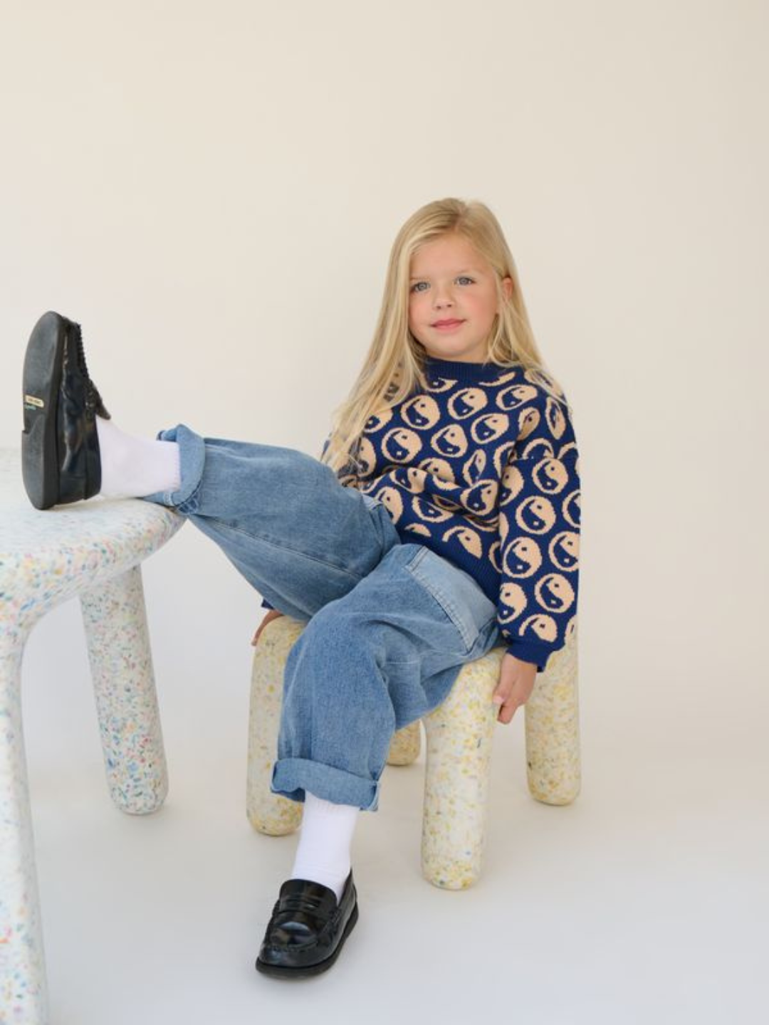 A young child with long blond hair sits on a stool, wearing the super-soft COSMOS SWEATER adorned with yin yangs, blue jeans, white socks, and black shoes, with one leg propped up on a nearby table.