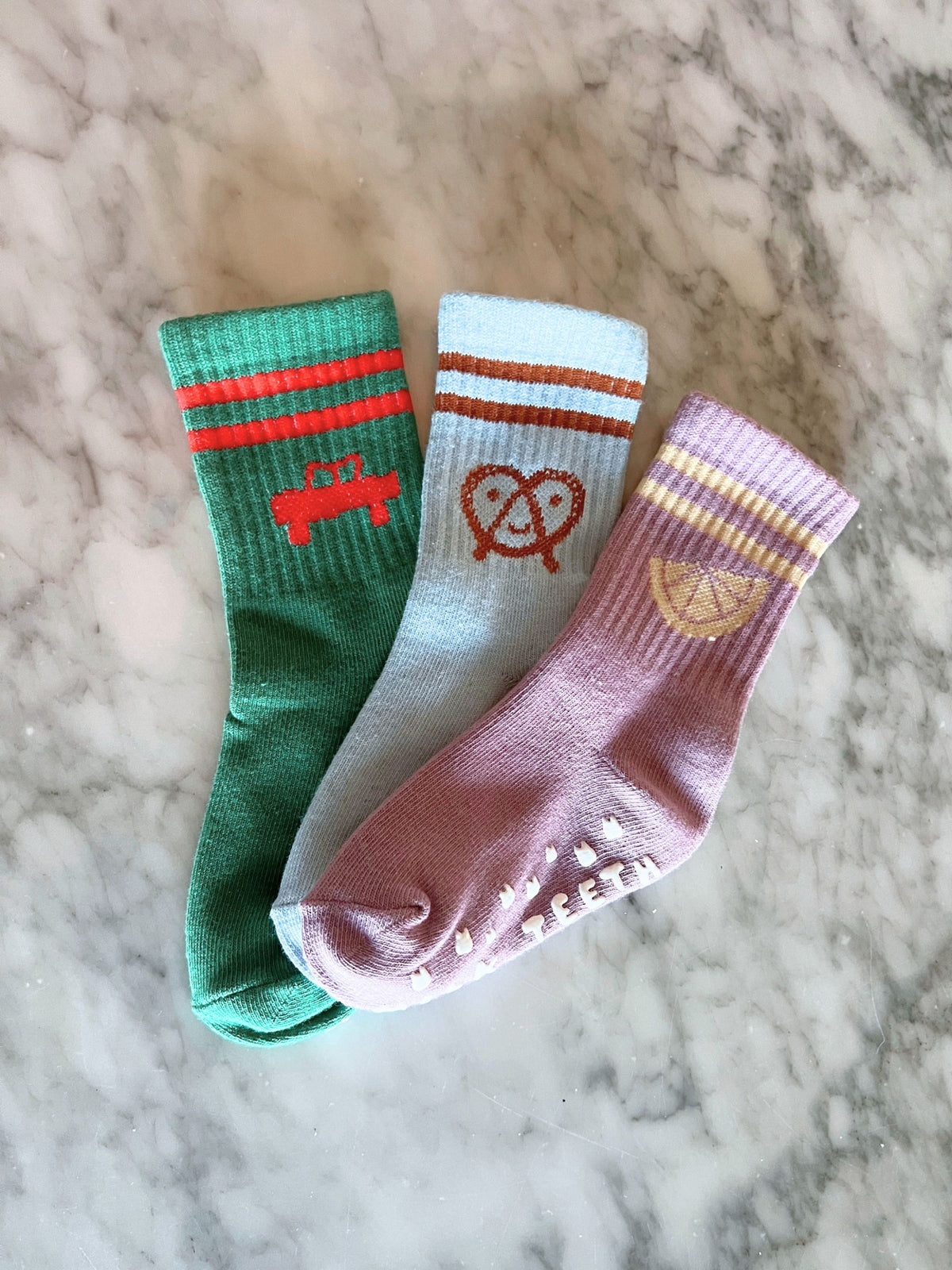 Three RIBBED GRIP SOCK pairs in green with a red car, light blue with a pretzel, and lavender with a lemon slice, all featuring anti-skid grips.