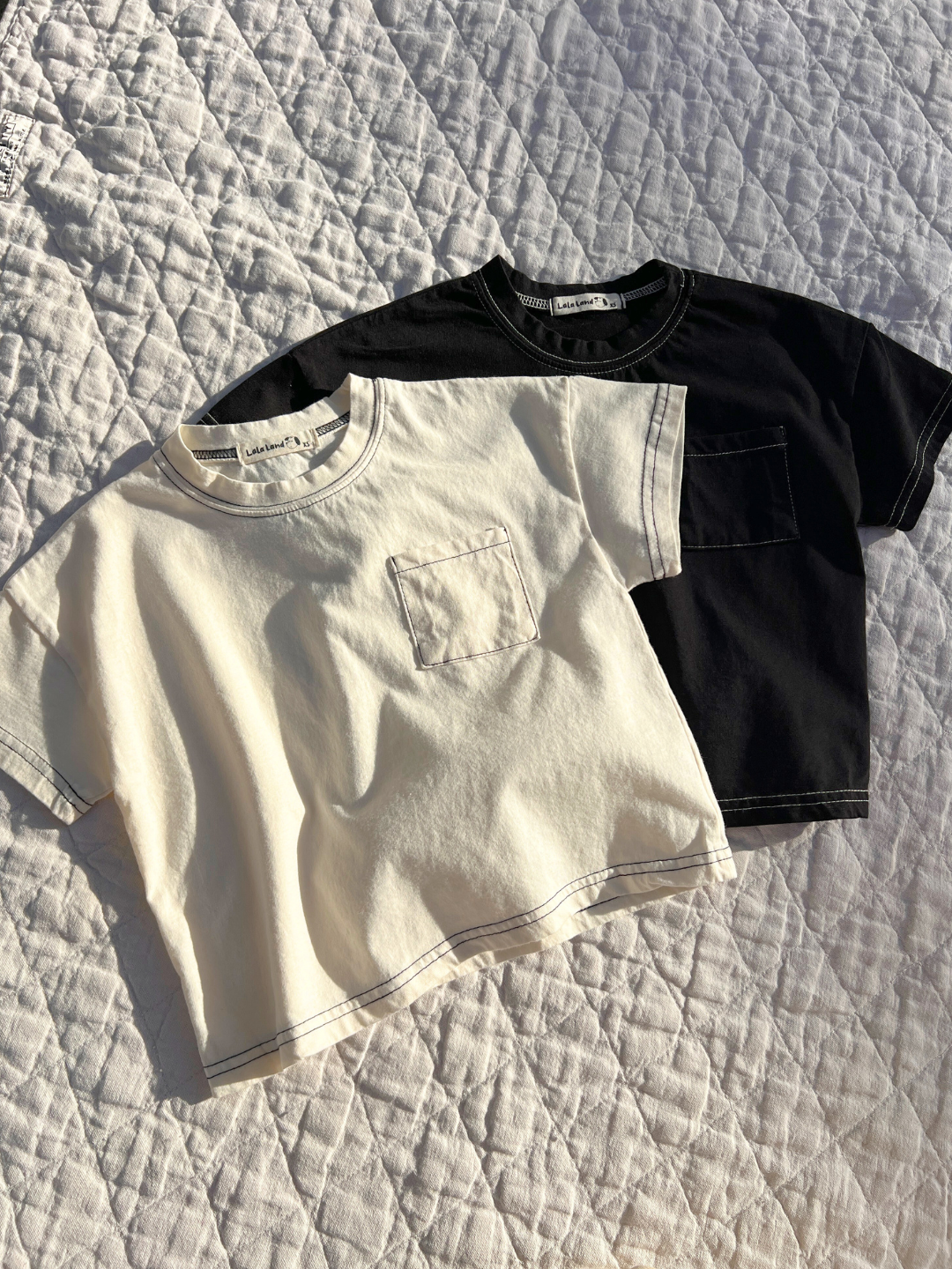 Ivory | Two of the kids' stitch tees are laid flat on a quilt in the sun