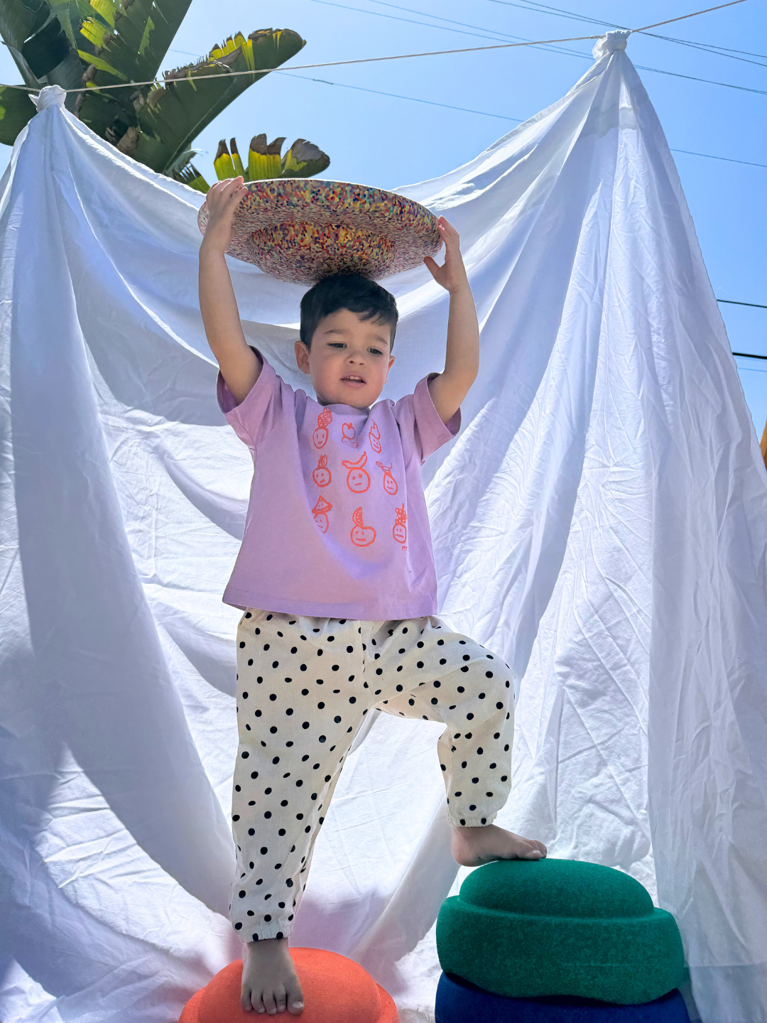 Child wearing the fruit faces tee in violet, with white pants with black polka dots. He stands on colorful play stepping stones and holds one over his head, in front of a white sheet backdrop.