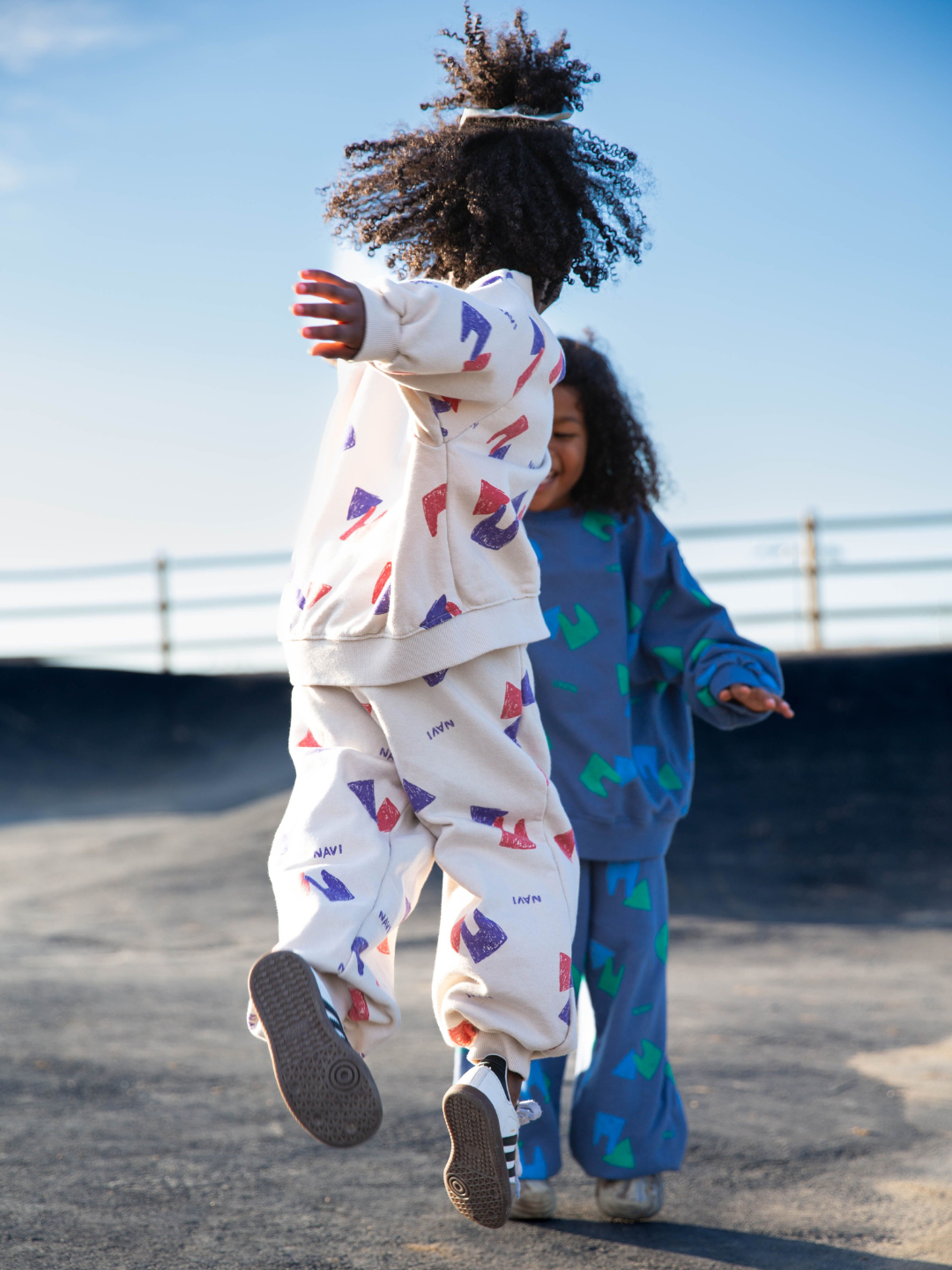 Oatmeal | Child wearing beige sweatpants with an all over pattern of red and purple shapes and the brand name Navi, with matching crewneck sweatshirt. She is jumping facing away from camera, at an outdoor skate park with blue sky. Another child is behind her in the same outfit in blue.