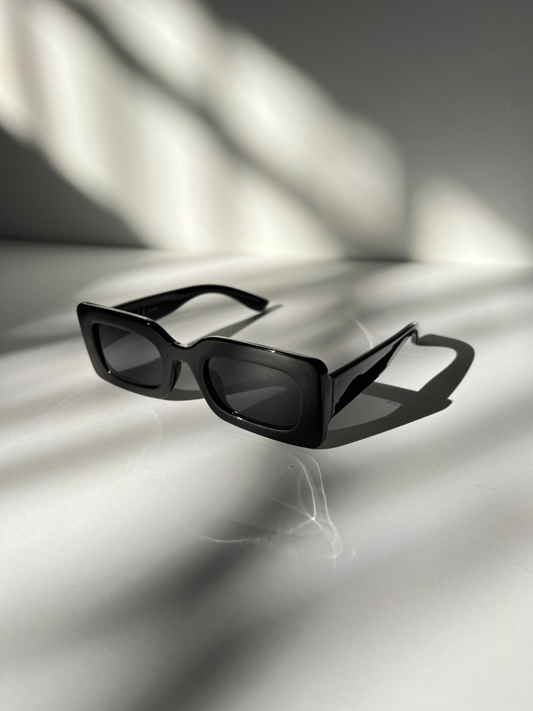 Kids rectangle sunglasses in black, on a grey background with areas of sunlight, creating shadows of the sunglasses.