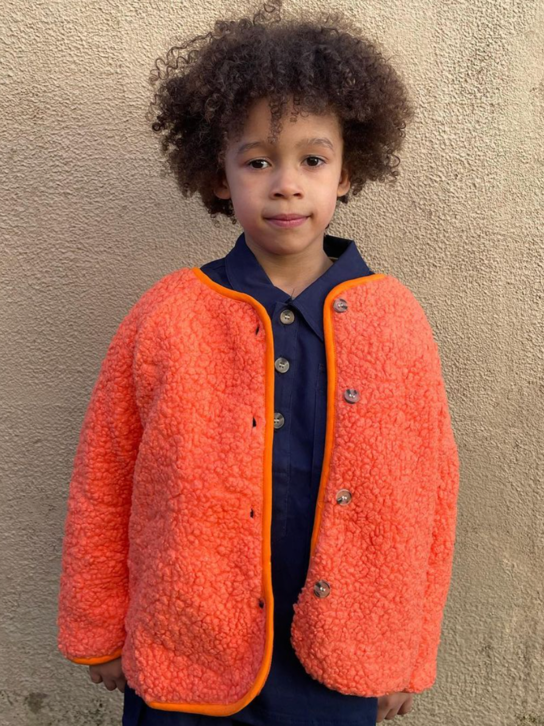 Tangerine | A child wearing a kids orange collarless fleece jacket with four brown buttons, worn over a navy blue shirt and pants, standing in front of a beige wall.