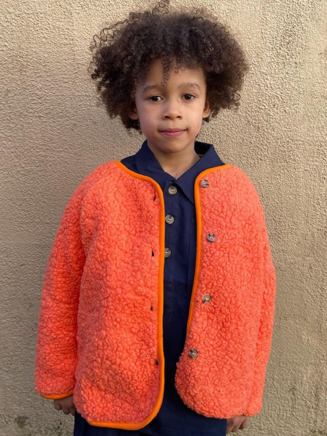 A child wearing a kids orange collarless fleece jacket with four brown buttons, worn over a navy blue shirt and pants, standing in front of a beige wall.