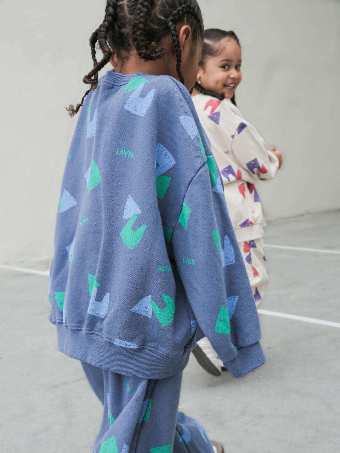 Child wearing blue crewneck sweatshirt with an all over pattern of green and blue shapes and the brand name Navi, with matching sweatpants.He is walking in front of a grey background.