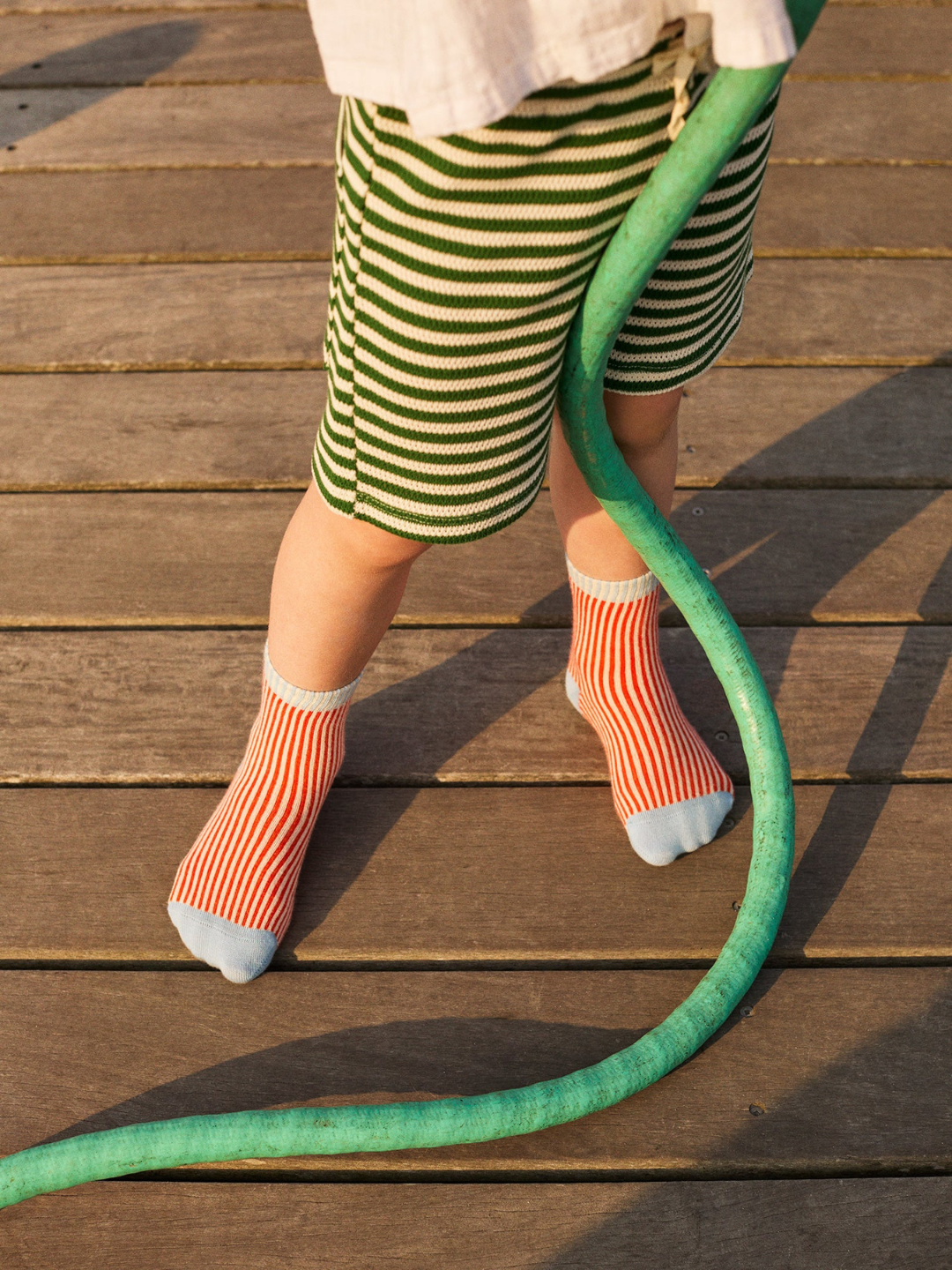 Kid wearing socks in narrow red and white vertical stripes with pale blue toe, heel, and green stripes at ankle cuff. Image is cropped below the waist showing a child wearing the socks, standing on a wooden deck, holding a green hose, and wearing green and cream striped shorts.