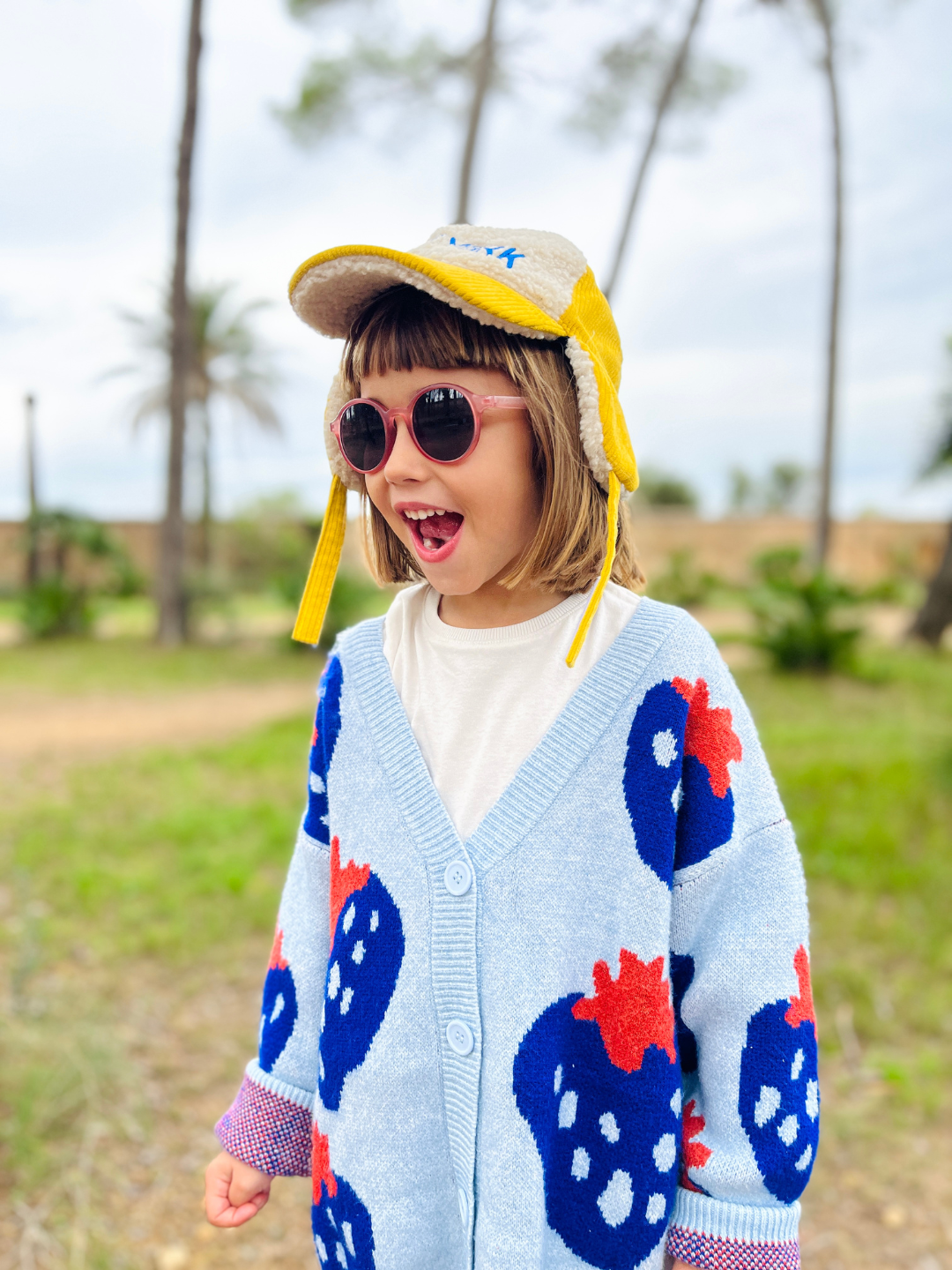 Sky | A girl wearing a kids v-neck cardigan in light blue with an all-over pattern of large blue strawberries with a red leaf, in an oversized fit. She is standing in a green park with palm trees in the distance, and wearing a white t-shirt, a yellow cap with ear flaps, and pink sunglasses.
