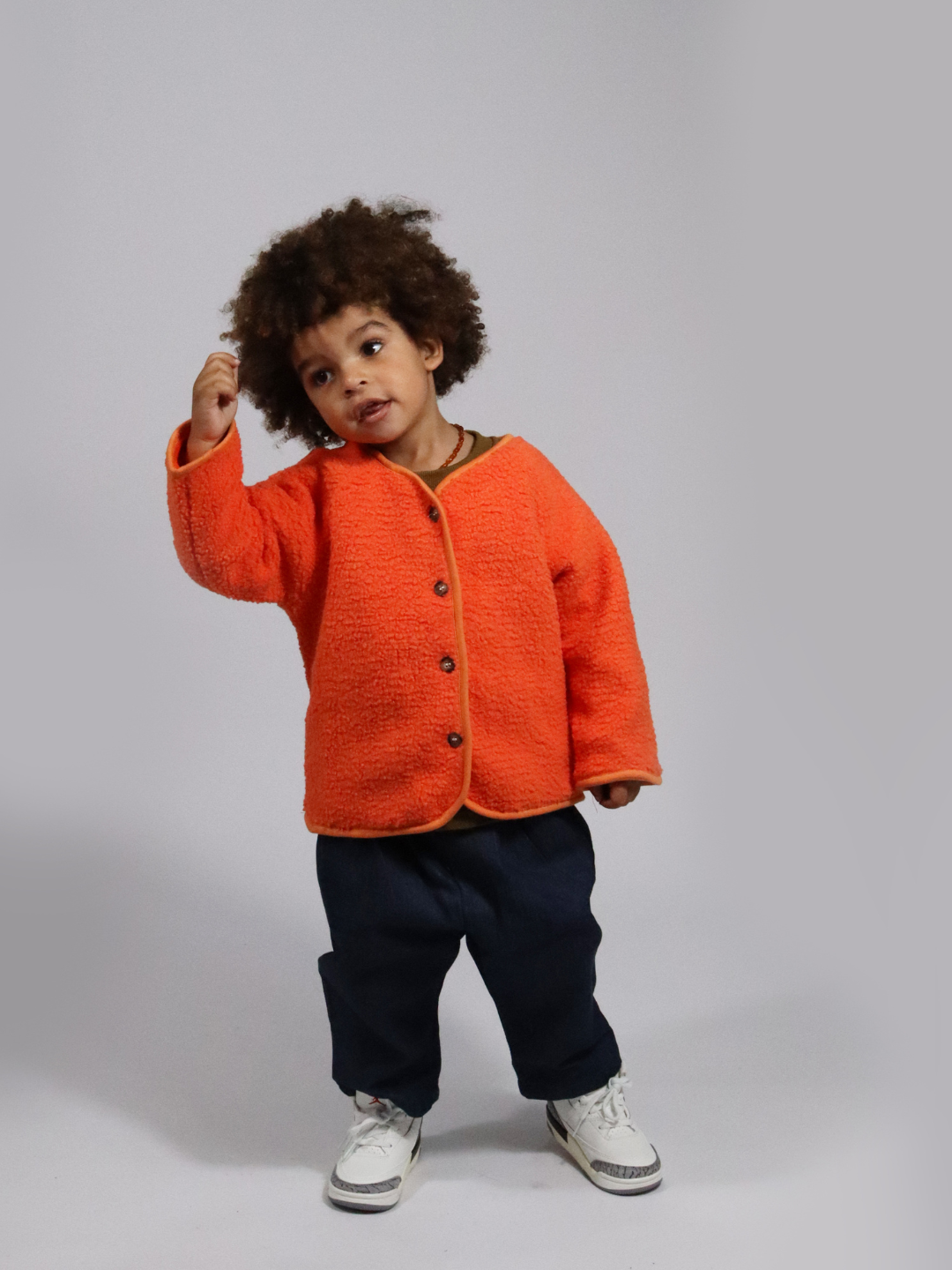 A toddler wearing an orange collarless fleece jacket with four brown buttons, with dark blue pants and white sneakers, standing on a light grey background.