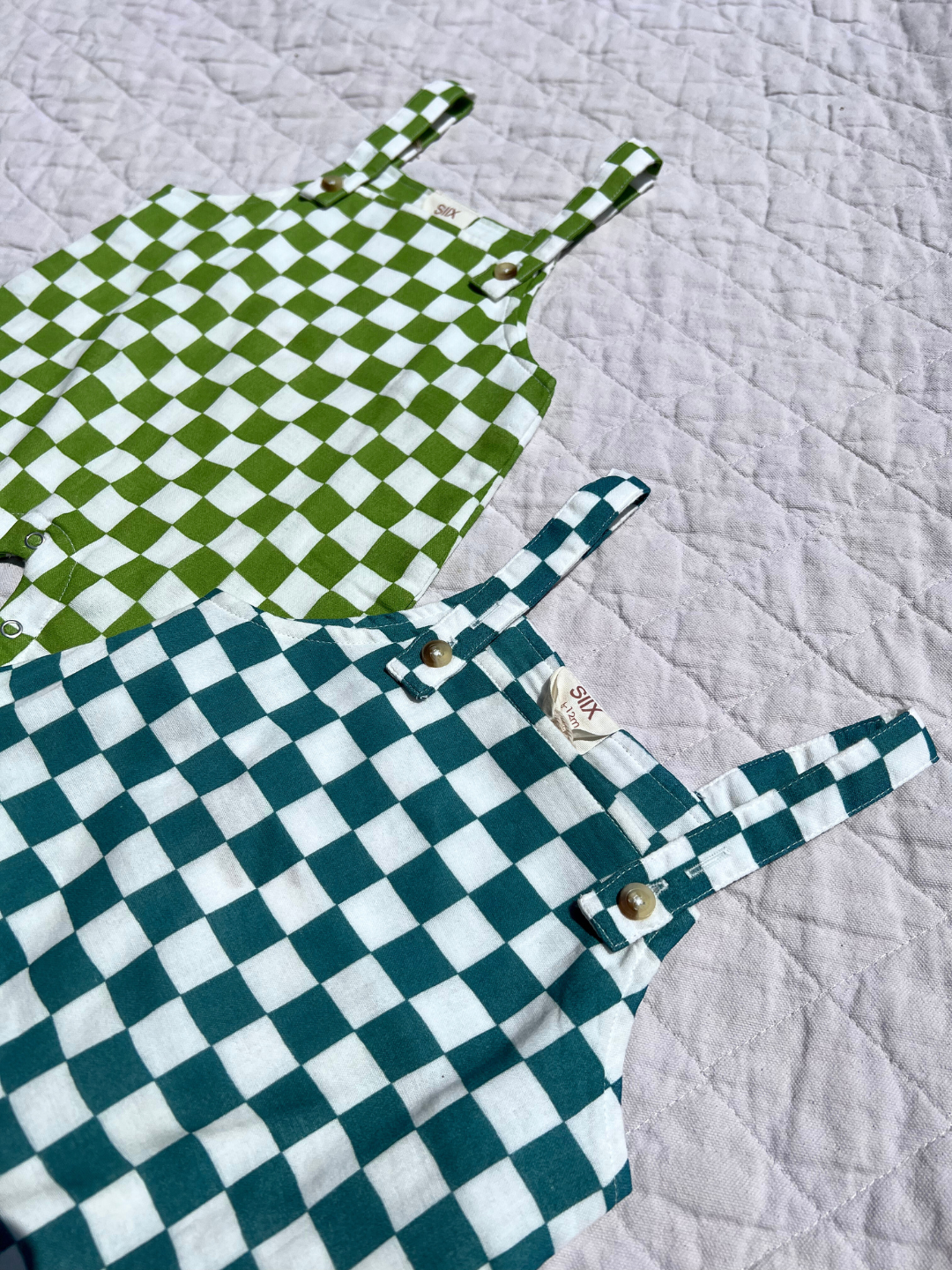 Close-up of two sets of kids' overalls showing buttons for adjusting straps; one in grass geen and white check, the other in aquamarine and white