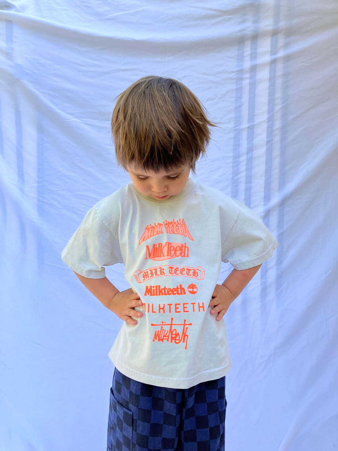 A child wearing a light grey t-shirt with Milk Teeth spelled out in various fonts in orange ink. He wears navy and black checkered pants and is standing in front of a white sheet backdrop.