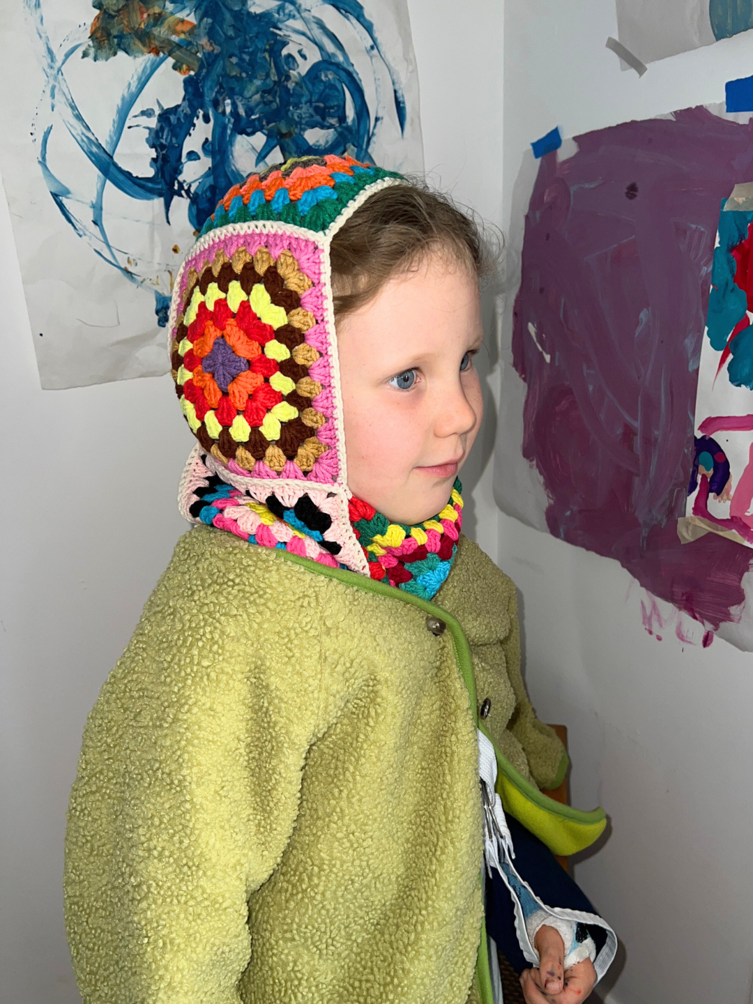 Child wearing the handmade colorful kids balaclava, with a green fleece jacket and her arm in a sling. She is smiling and standing in front of kids paintings on a white wall.