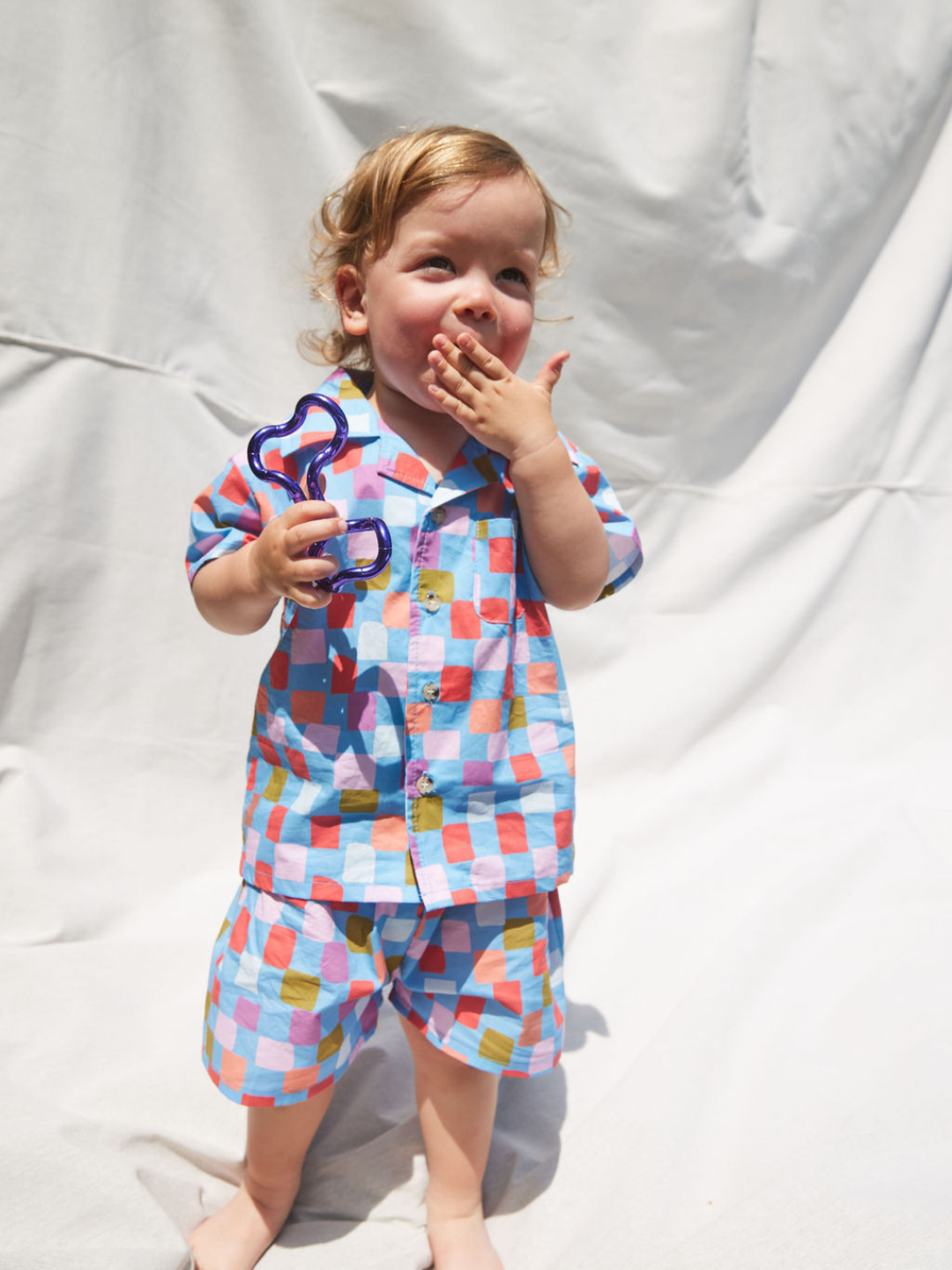 A toddler wearing a kids' shirt and shorts set in a pattern of red, orange, pink, lilac and green squares on a blue background