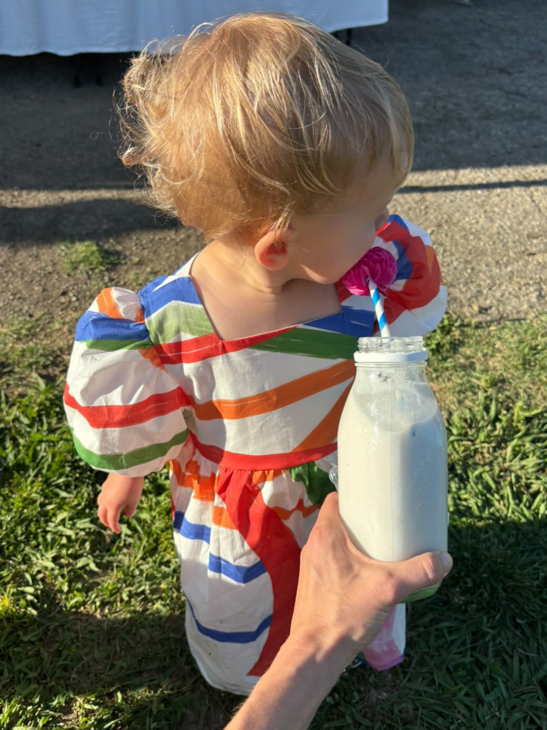 Colors | A toddler wearing the kids Glow Dress in colors featuring paintbrush-style lines of bold color all over a white cotton dress. She is drinking milk from a bottle with a straw, held by an adult's hand, and standing on grass looking away from camera.