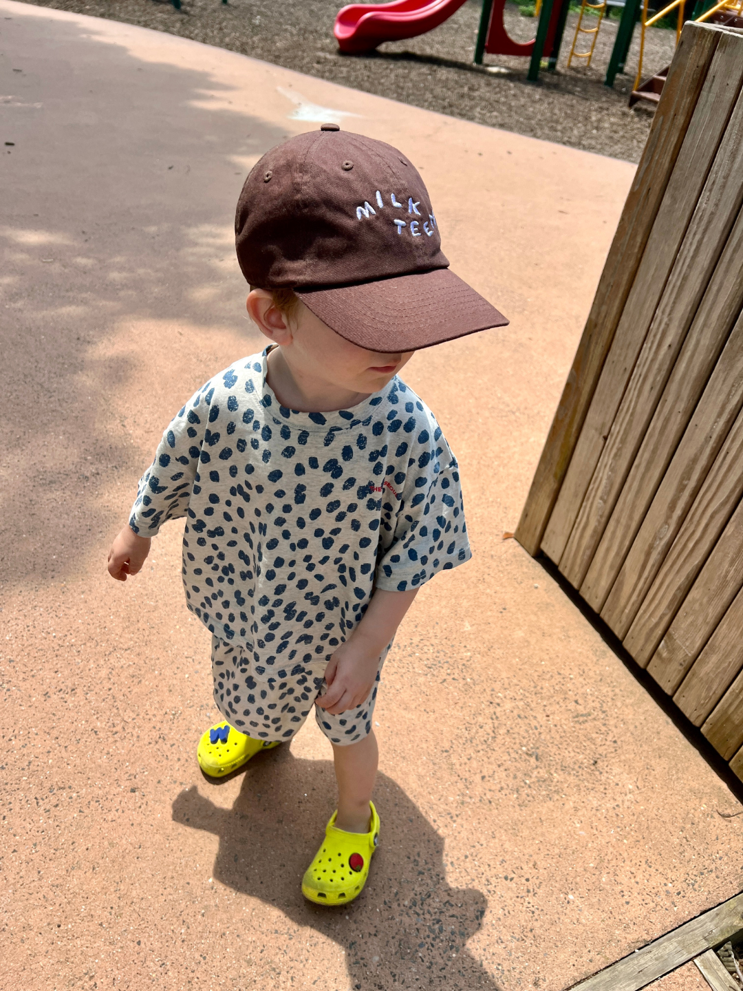 A young child wearing a brown cap labeled "milk tea," a SPOTTED TEE in white with blue polka dots, and bright yellow crocs stands on a playground. The soft, orange ground complements the play area in the background, adding charm to the scene. This adorable cotton jersey top is from a popular Korean kids' brand.