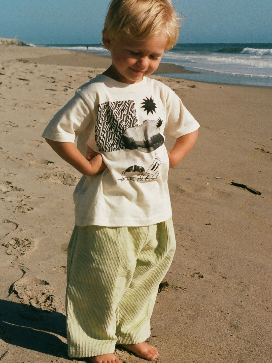 A child on a beach wearing a kids' tee shirt in cream, printed in black with stars, zigzags, a PBJ sandwich, a toaster and the words Milk Teeth