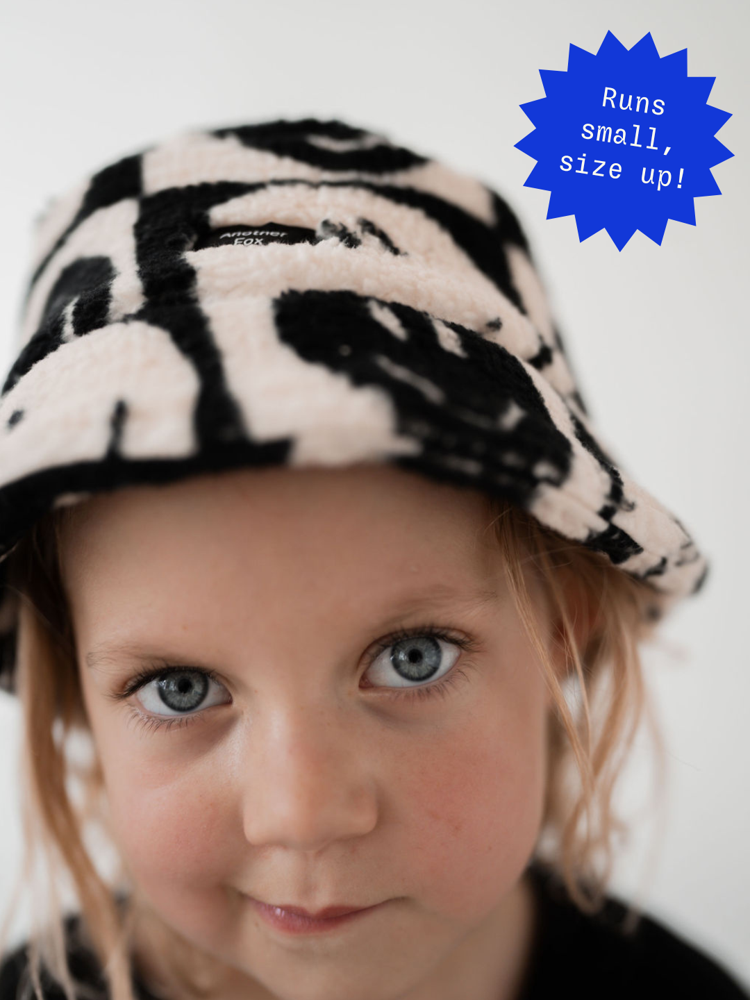 A young child with blue eyes wears a FACE CHECKER BUCKET HAT from our latest clothing new arrivals, looking into the camera with a neutral expression. A blue badge on the image says that the hat runs small, and to size up.