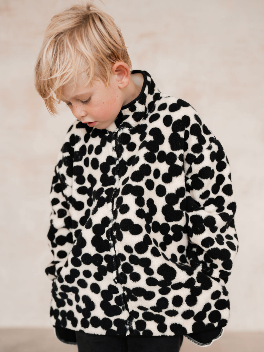 A young child with blond hair wearing the SPOT FUNNEL COAT—an adorable black and white polka dot jacket—looks down, hands in pockets. Shop all clothing items to find similar styles and check prices for new arrivals.