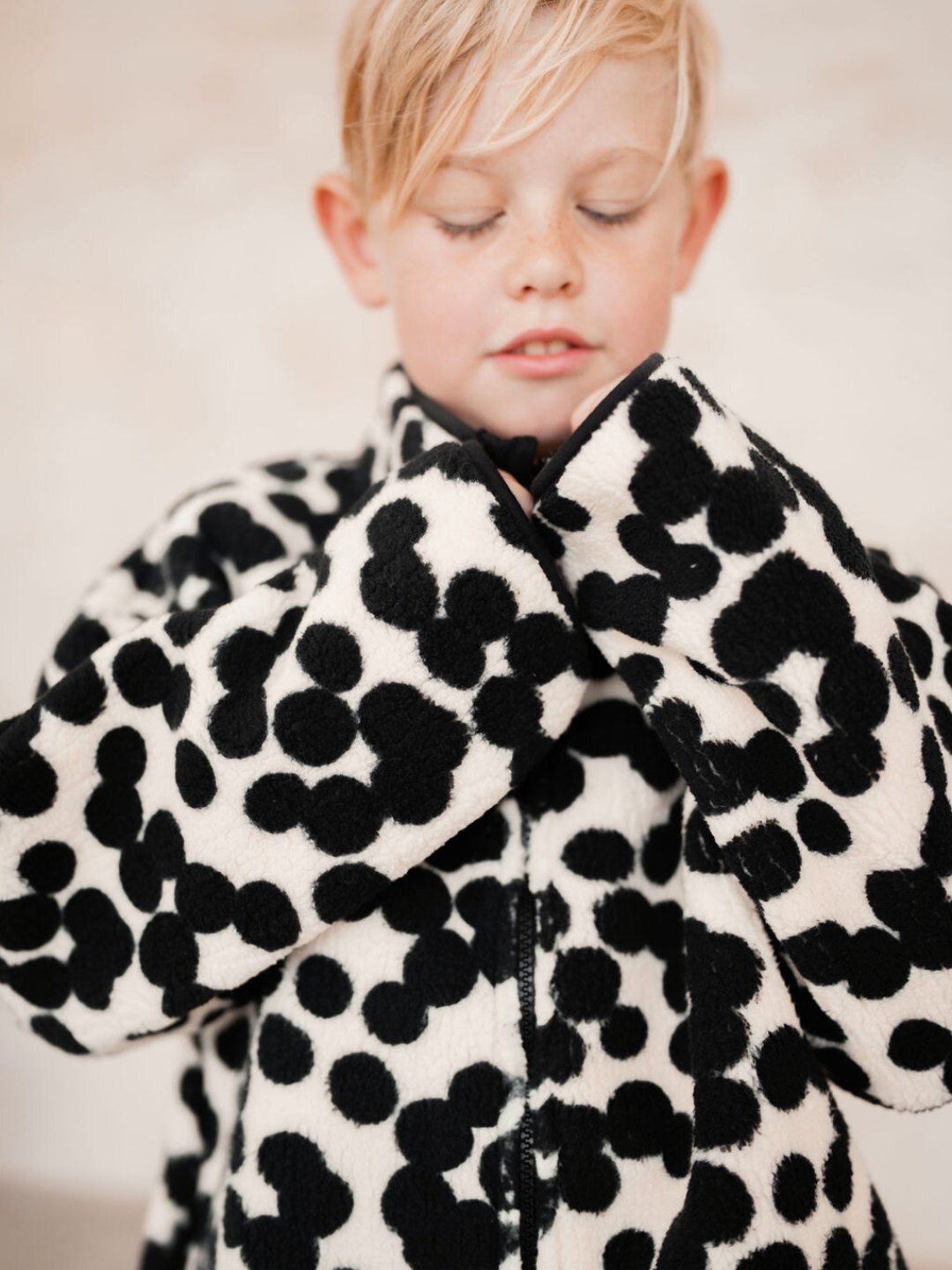 A baby with closed eyes is wearing the SPOT FUNNEL COAT, a black and white polka-dotted jacket. Shop all clothing to discover similar adorable styles.