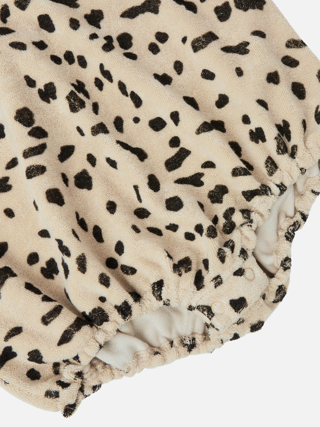 Close-up of the DALMATIAN TERRY BODYSUIT, featuring a beige fabric with an irregular black spot pattern reminiscent of dalmatian spots. The ruched, elastic edges suggest it is a comfortable and stylish addition to the new arrivals collection for infants.