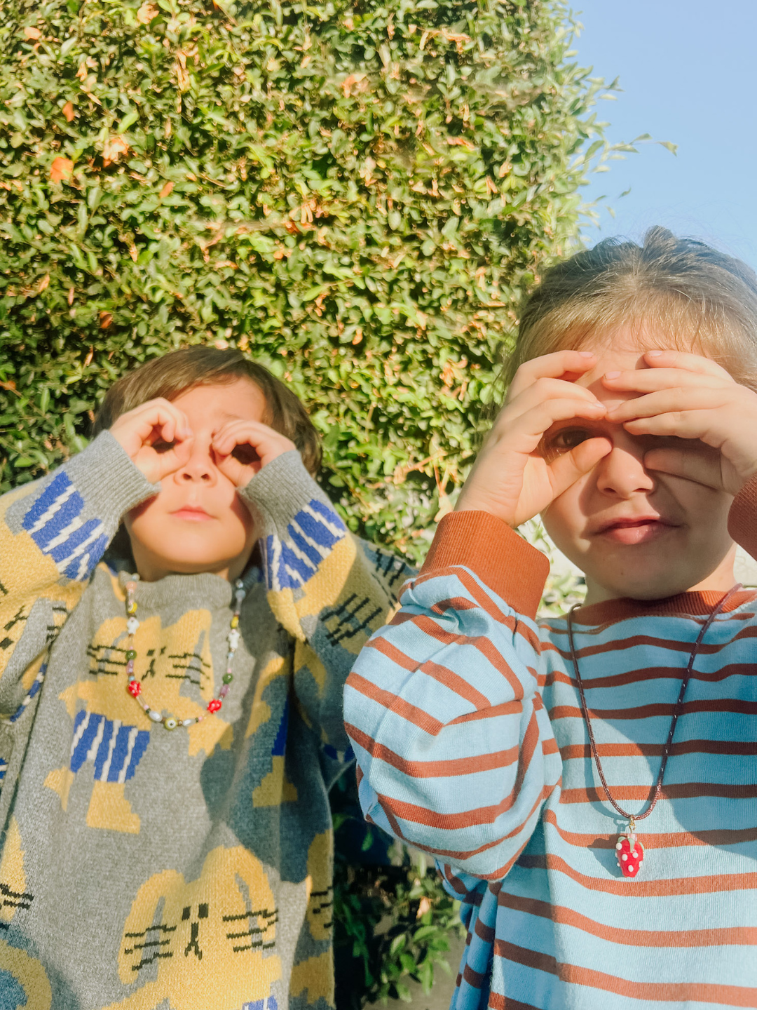 Two children stand outside before greenery, making pretend glasses with their hands. Both wear patterned sweaters, beaded necklaces, and the playful FRAGOLA NECKLACE from Meyous jewelry.