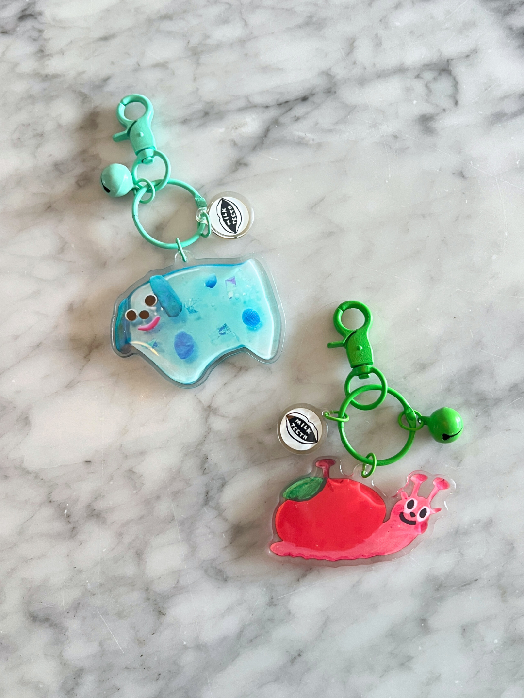 Dog | Two SOUVENIR KEYCHAINs by Katie Kimmel—a blue elephant and a pink snail on a red apple—sit on a marble surface. Each has green clips and round charms, making them playful bag accessories.