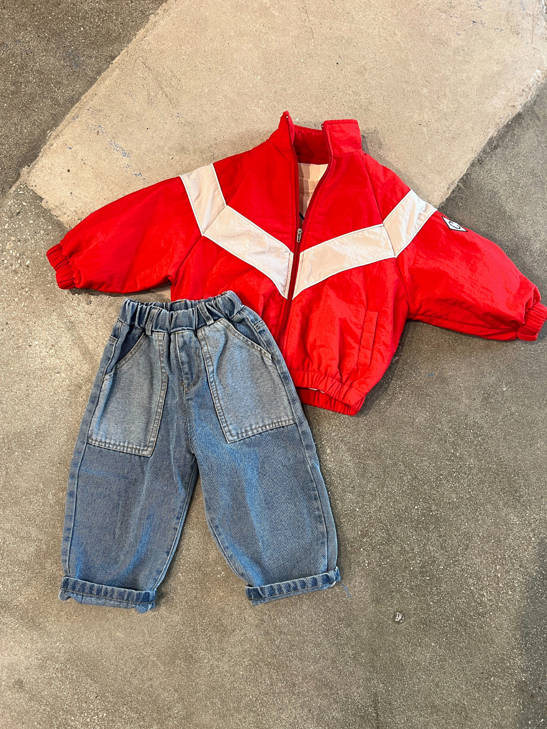 Red | Children's outfit laying on a concrete surface, featuring a WARM-UP JACKET with white chevron stripes from a Korean label and a pair of blue denim jeans with an elastic waistband and rolled-up cuffs.