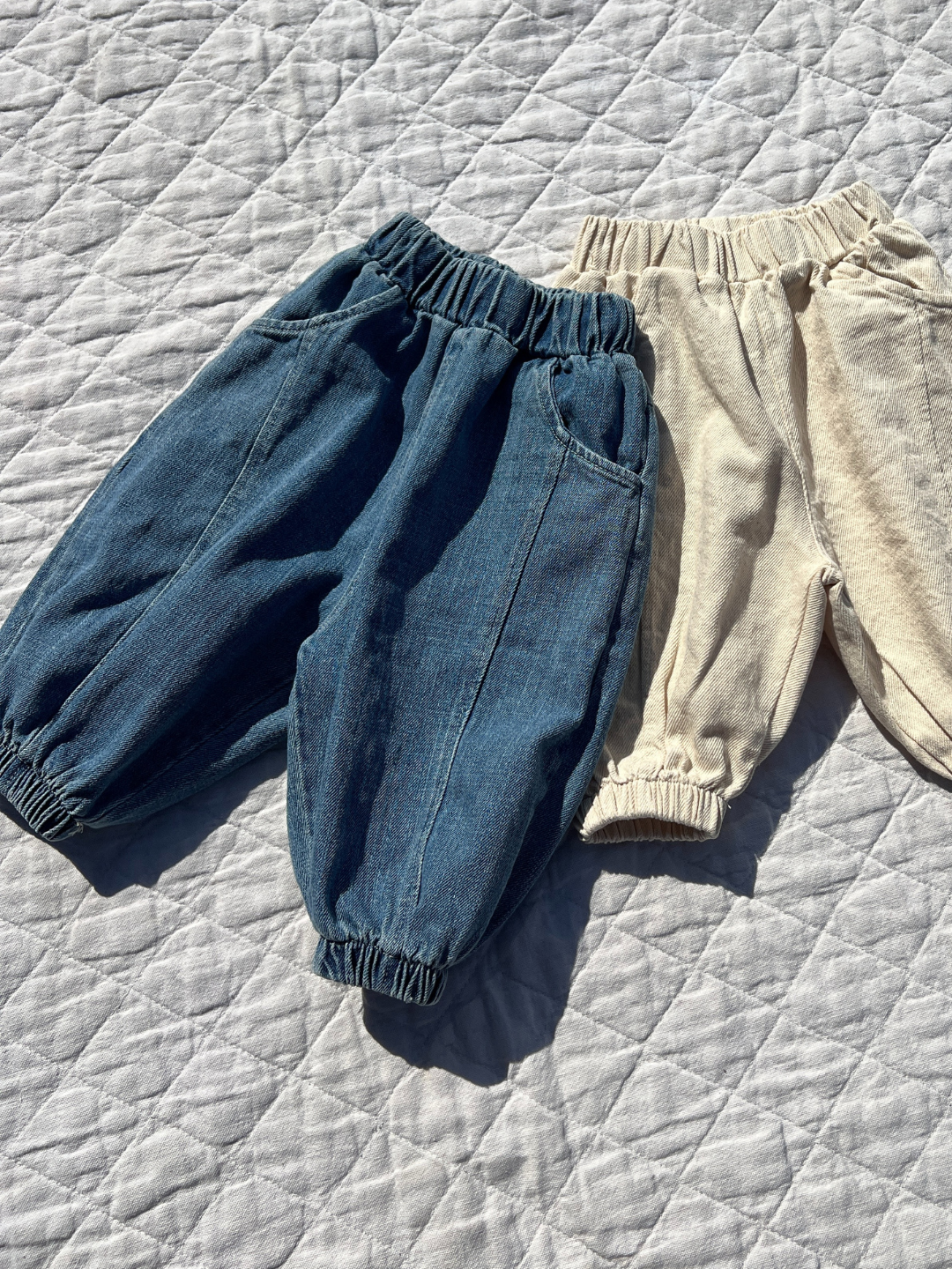 Denim | Two pairs of children's pants rest on a quilted surface: one pair is Valu Baby's denim blue BALLOON JEAN for babies, while the other is light beige with a jogger-style fit, both highlighting Valu Baby's dedication to comfort and style.