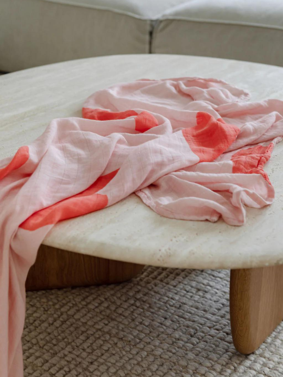 A light pink and coral fabric, reminiscent of the ORGANIC BAMBOO SWADDLE WRAP, is draped over a white, oval-shaped coffee table with wooden legs, set on a textured beige carpet.