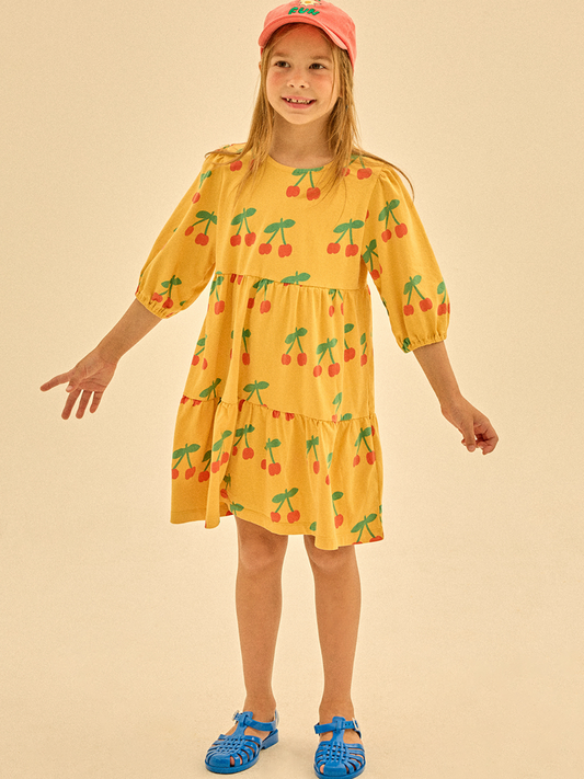 Second image of The CHERRY RUFFLE DRESS is a yellow children's dress with three-quarter sleeves, a tiered skirt, and a cherry pattern featuring red cherries and green stems on a white background.