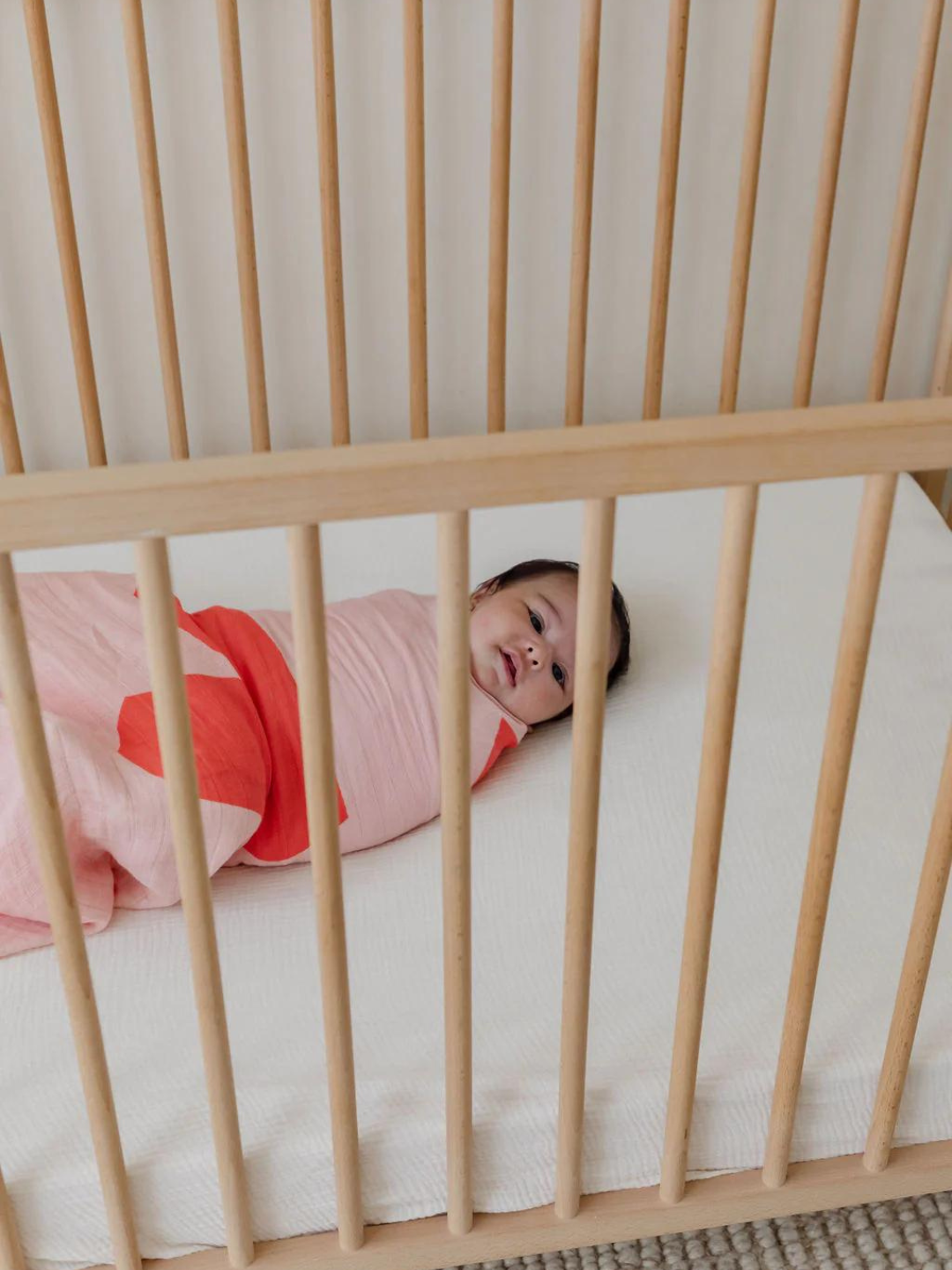 A baby wrapped in an ORGANIC BAMBOO SWADDLE WRAP lies in a wooden crib, staring up.