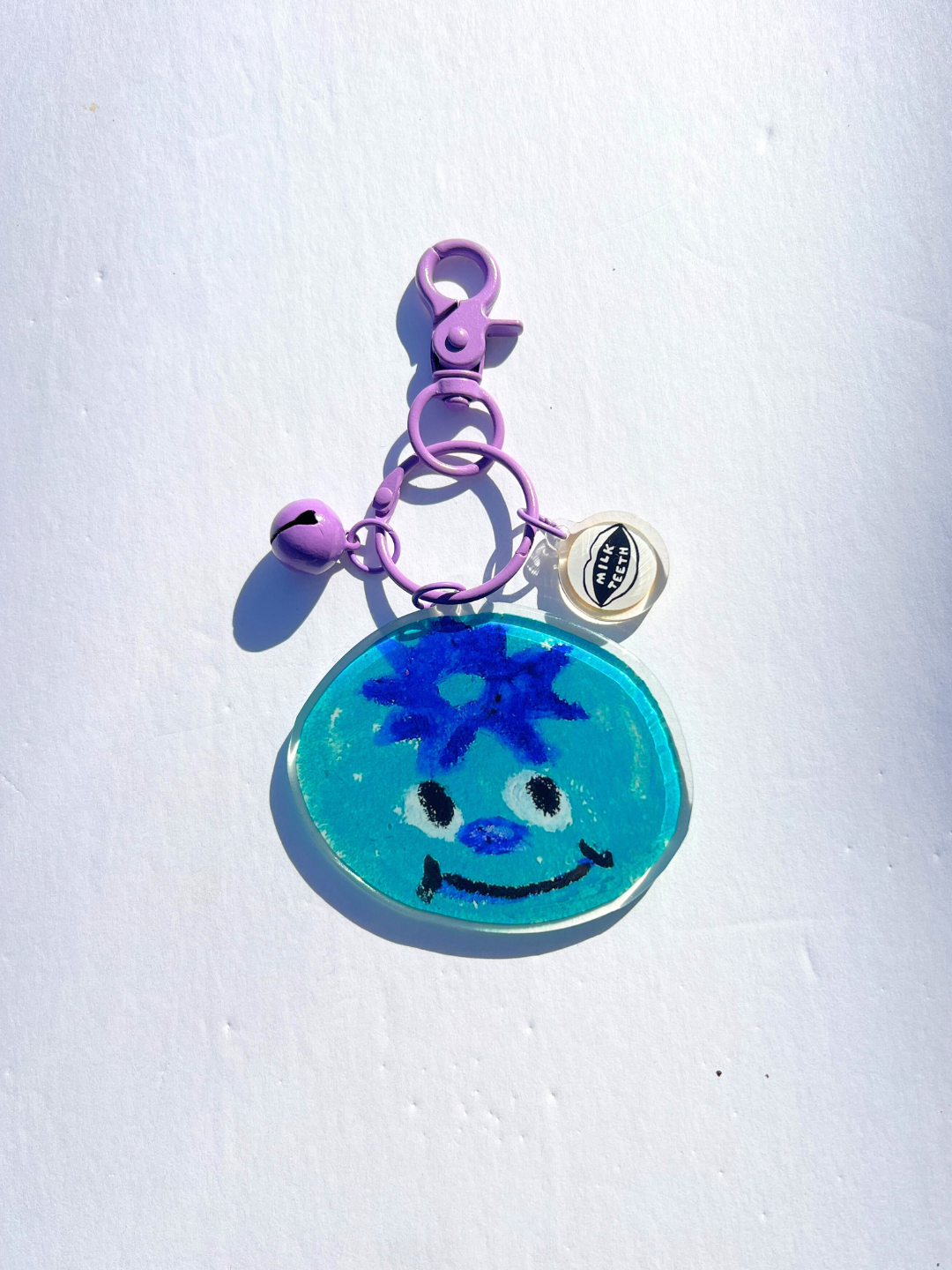 The SOUVENIR KEYCHAIN is a blue smiling face charm with a purple clip, small bell, and round white tag—perfect for accessorizing bags. Photographed on a white background.