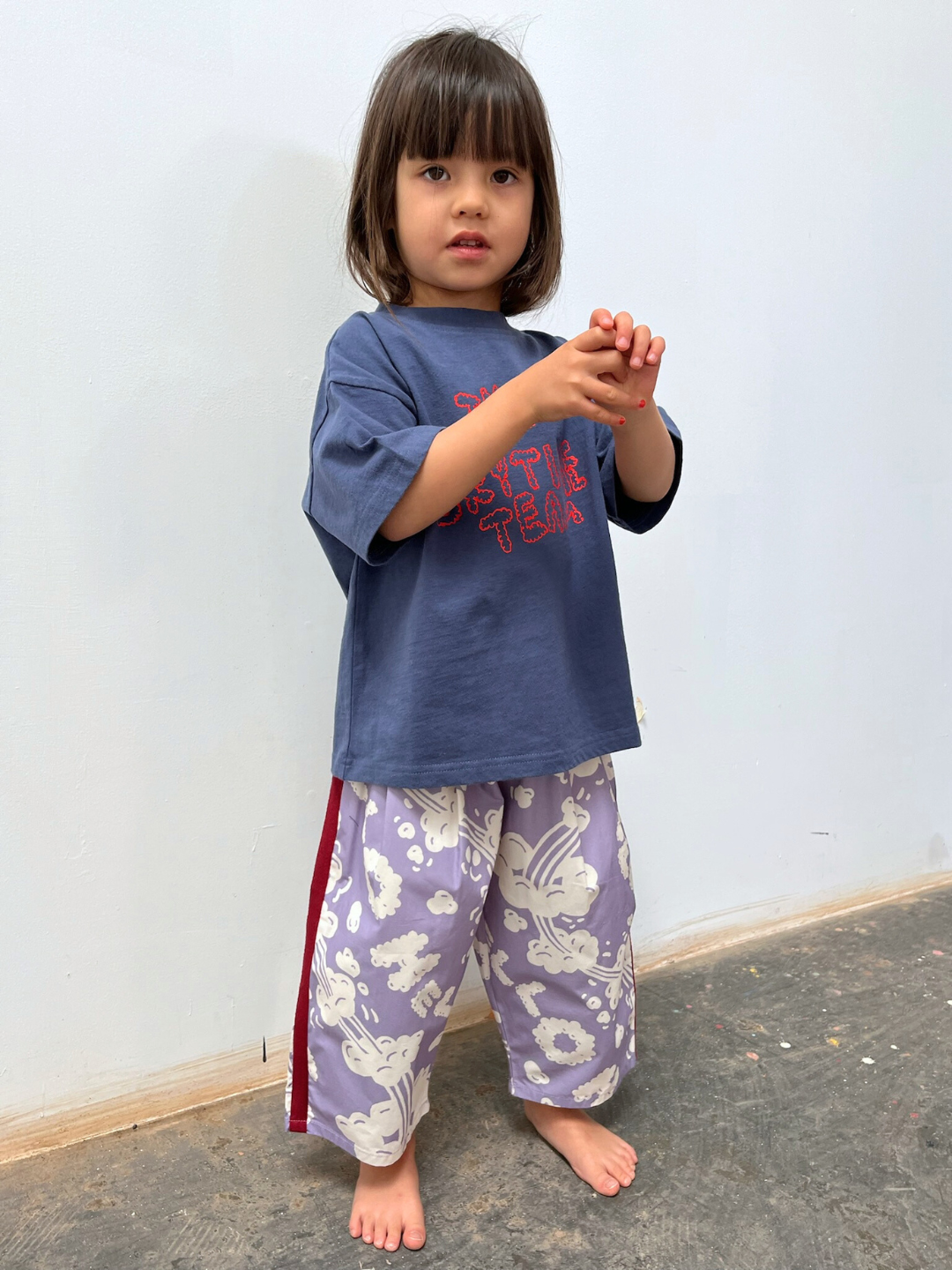Purple | A young child with shoulder-length hair stands barefoot on a concrete floor, wearing loose lavender SKYWRITING PANT and a blue oversized T-shirt against a plain light background.