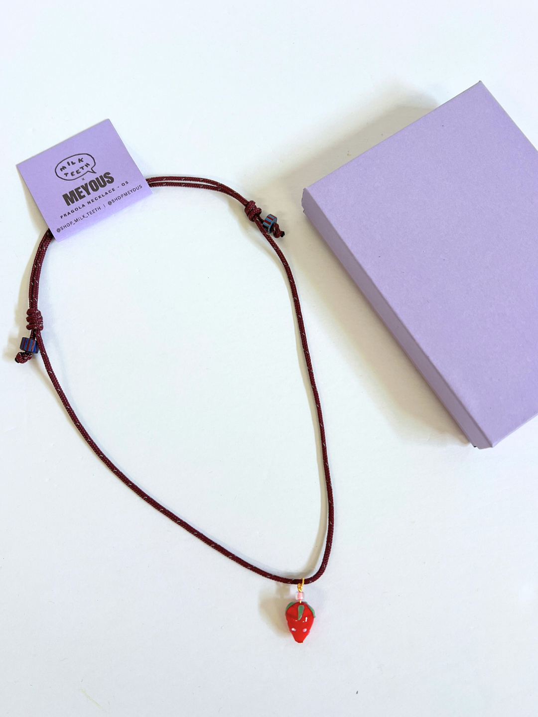 The FRAGOLA NECKLACE, featuring a brown cord and handmade glass strawberry charm, is displayed beside a closed lavender box on a white surface, highlighting the distinctive style of Meyous jewelry.